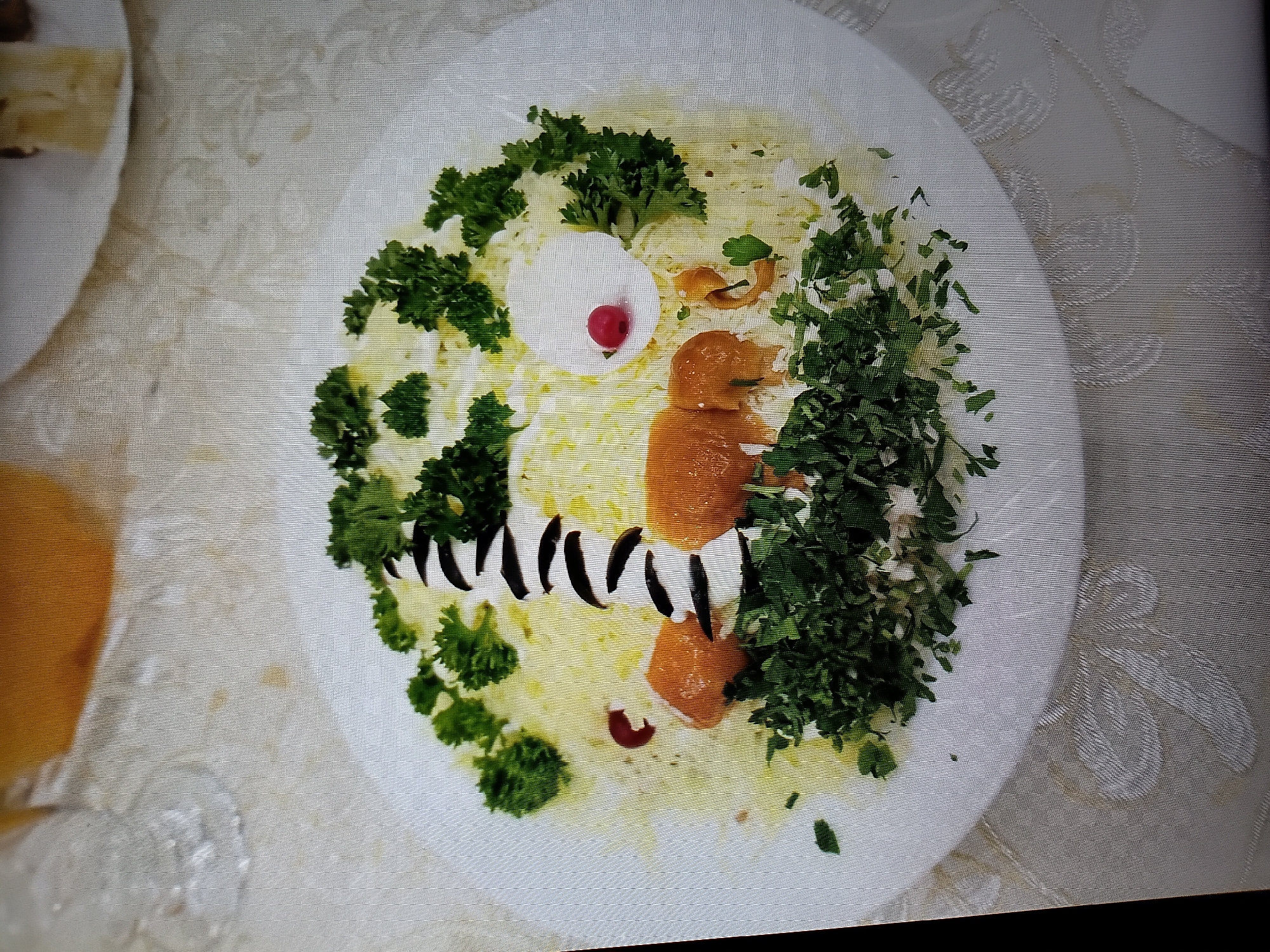 The image shows a close-up, high-angle shot of a creatively plated salad on a white, circular dish. The salad is arranged to resemble a lighthouse, with a base of shredded white ingredients (likely potato or cheese) and a cylindrical structure made of what appears to be a white creamy substance, possibly mayonnaise or sour cream, decorated with black olive slices to create stripes. At the top of the lighthouse structure, there is a white circle topped with a single red berry, resembling a beacon. The entire dish is garnished with chopped fresh parsley, giving it a green, leafy appearance. There are also pieces of what look like cooked shrimp or fish, in an orange hue, positioned around the base of the lighthouse. The dish is placed on a tablecloth with a subtle, patterned texture. The background is blurred, but appears to be an indoor setting with other dishes and glassware visible, suggesting a restaurant or dining environment. There is no indication of the city of Tiraspol, Moldova, in the image itself.