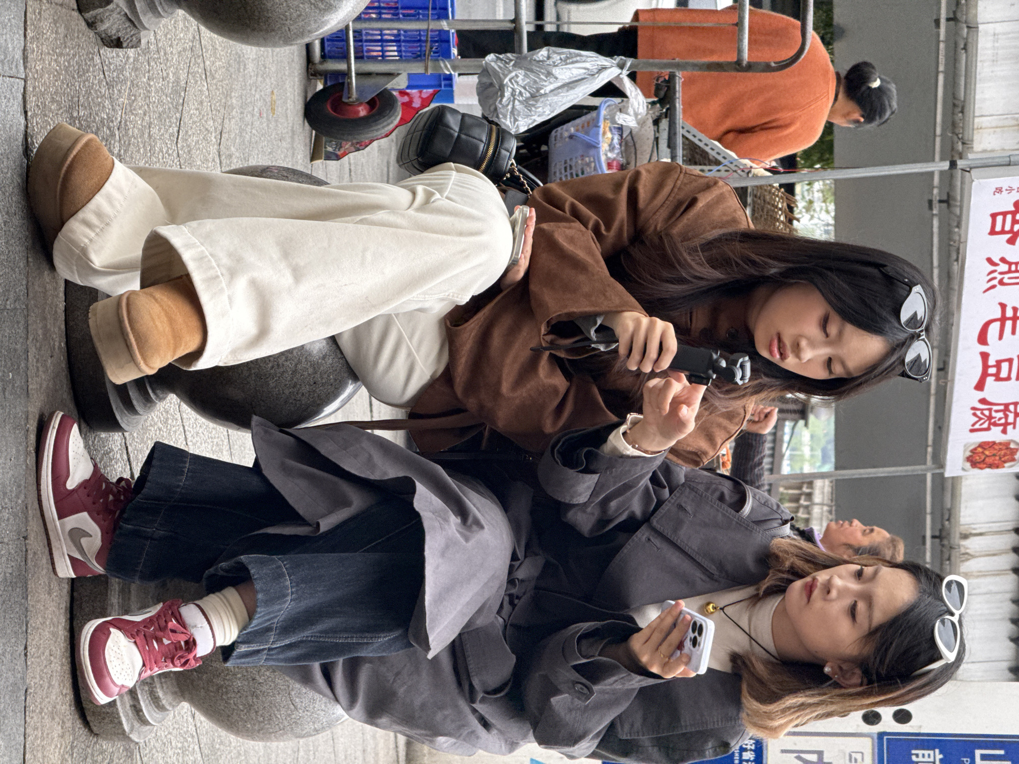 Two young East Asian females are seated on cylindrical gray stone bollards in a public area of Yuzhong District, China. The female on the left wears a brown jacket, white wide-leg pants, and brown platform slippers, with dark sunglasses resting on her long dark hair. She holds a black handheld device, possibly a portable fan or small gimbal, in her right hand, while her left hand is raised in a gesture. A small black quilted shoulder bag is visible on her left side. The female on the right wears a dark gray trench coat over a white turtleneck, dark blue wide-leg jeans, and red and white sneakers, with white cat-eye sunglasses resting on her brown ombre hair. She holds a light-colored smartphone with a purple case, engaging with its screen. Both individuals are focused on their devices.
In the background, an older East Asian woman in an orange sweater with her hair in a bun is partially visible, facing away from the camera. To the left, a metal structure with a blue plastic basket containing various items and a silver plastic bag hanging is present, along with a red-wheeled cart. Directly behind the seated females, a white sign with red Chinese characters, likely "酱黑毛豆腐" (fermented black hairy tofu), is partially visible. To the right, a blue sign with white text, including some Chinese characters and English, is partially visible. The ground is paved with gray stone slabs, characteristic of an urban street or market setting.