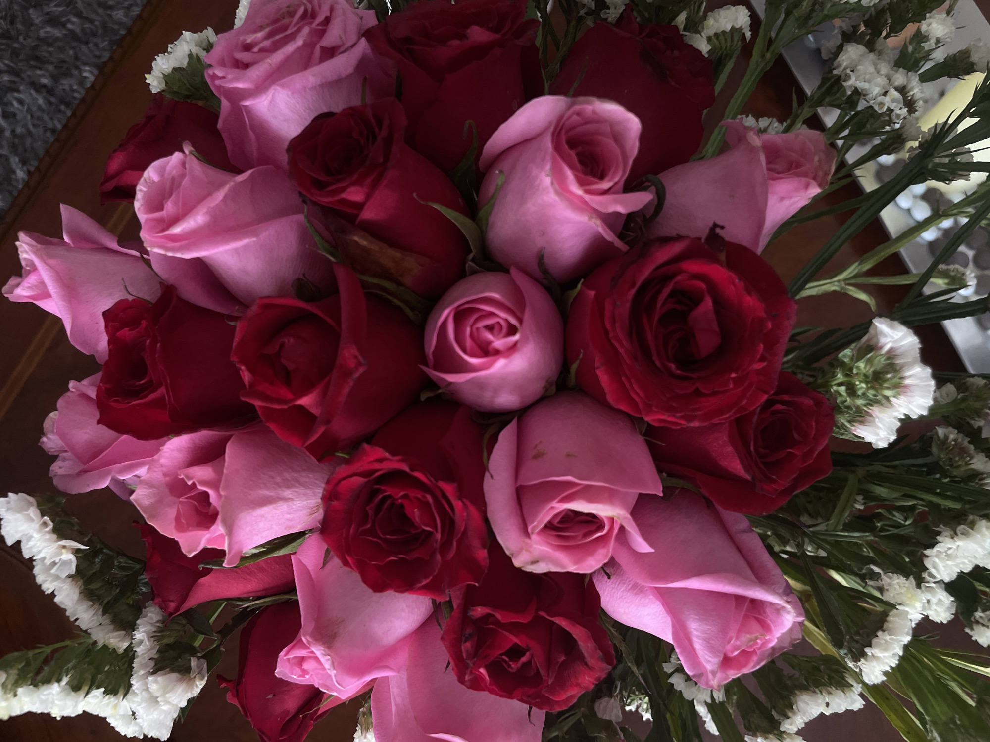 A bouquet of roses is displayed, featuring a mix of pink and red roses. The roses are tightly packed together, creating a dense arrangement. Small white flowers are interspersed among the roses, adding to the fullness of the bouquet. The bouquet is set on a dark wooden surface. The location is not evident from the image.