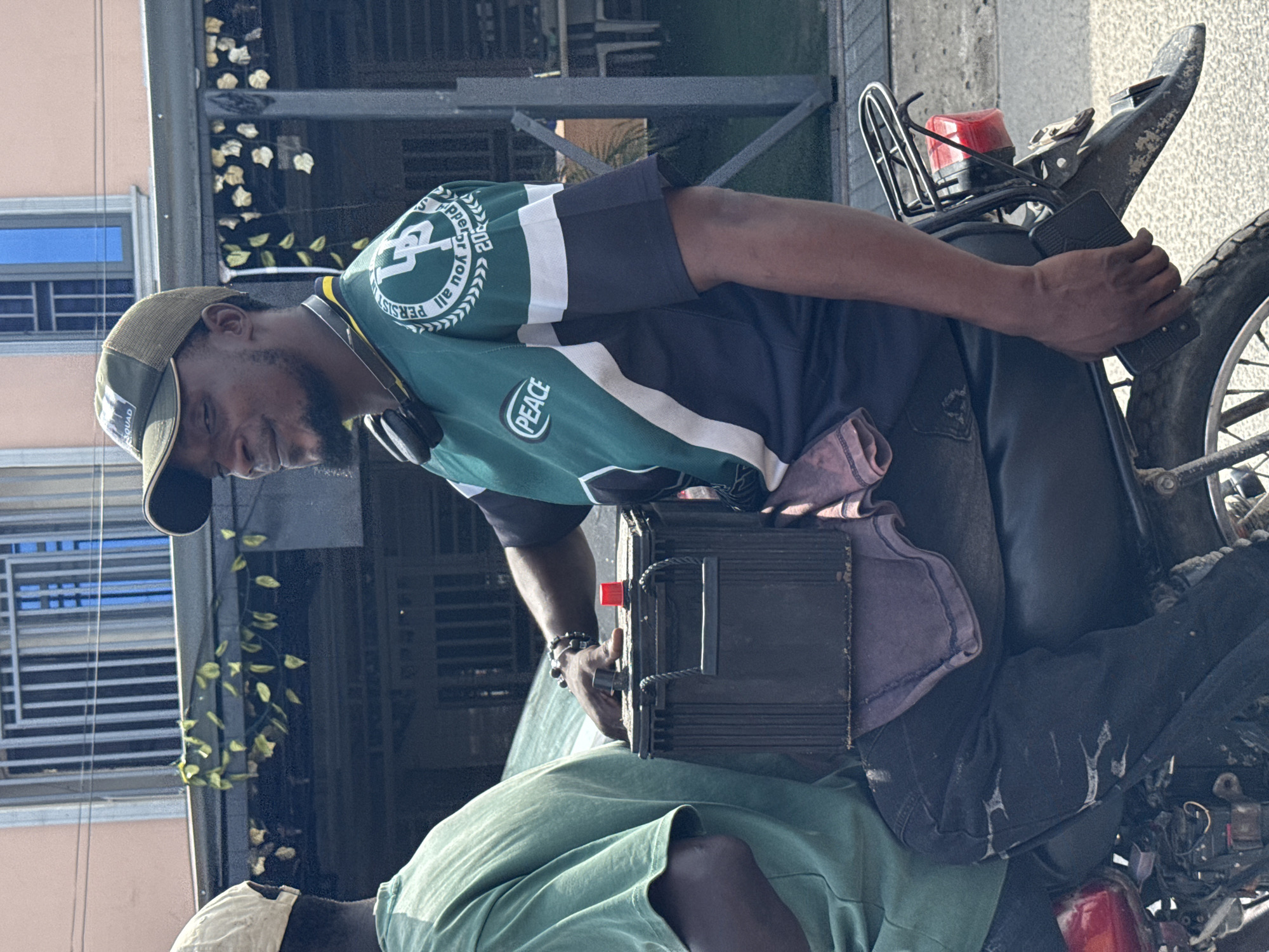 Ado, Nigeria. A man with a cap and headphones is wearing a green and white shirt that says "Peace." He is bent over, holding a black rectangular object with his left hand while seated on a motorcycle. The motorcycle has a red taillight and a dusty rear tire. The background shows a multi-story building with a window and a black metal railing with floral decoration. Another man is sitting on the motorbike with a green shirt.