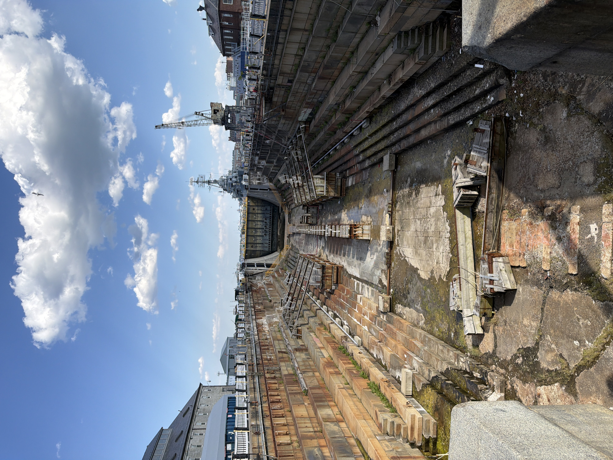 The image depicts a dry dock in Boston, United States of America. The dry dock is a large, rectangular structure with stepped sides. The walls of the dock are made of concrete and stone. The bottom of the dock is concrete. There are several large ships in the dock, and a crane. The sky is blue with white clouds. Some buildings are visible on the upper edges of the dry dock.