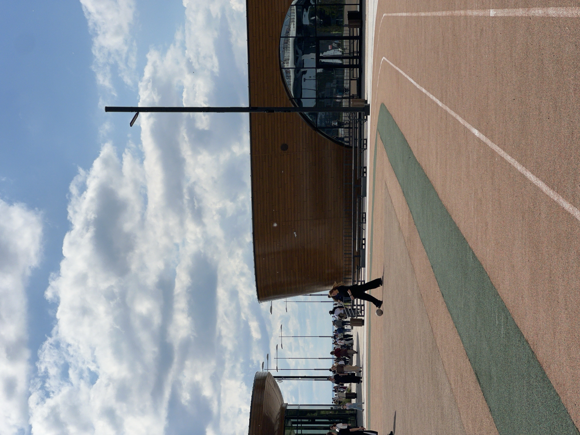 The image depicts a public outdoor space with a modern architectural design, featuring two curved buildings with wooden facades and large glass windows. A paved area in the foreground has a running track marked with white lines and a green strip. In the mid-ground, a woman in dark clothing is walking with a soccer ball. A crowd of people is visible in the background, walking along a pathway in front of the buildings. Several lampposts are scattered throughout the scene. The sky is partly cloudy. This scene is likely located in the area of the Krestovsky Island in Saint Petersburg, Russia, specifically near the FLC Zenit Arena complex, known for its contemporary design and recreational facilities.