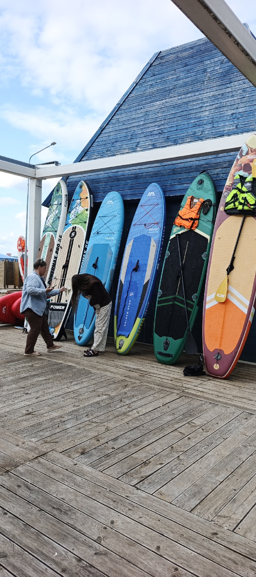 The image shows a collection of stand-up paddleboards (SUPs) lined up against a dark blue wooden building. Two people are visible near the paddleboards. One person, wearing a blue shirt and brown pants, appears to be holding a phone. The other person, with long dark hair, is bending over and seems to be cleaning or adjusting something.
The SUPs are of various colors and designs, with some featuring branding like "POWERMAX PRO 116," "AM," "VAPOR," and "BEAST." One paddleboard has an orange life vest and sandals placed on it. A paddle with "ALOHA" written on it is leaning against another board.
The scene is set on a wooden boardwalk, which is made of weathered grey planks arranged in a diagonal pattern. In the background, the upper part of the blue building with a sloped roof is visible against a sky with scattered clouds. A streetlamp is also present to the left of the building.
The overall impression is a casual outdoor setting, possibly a rental or storage area for water sports equipment. The lighting suggests it is daytime, with cloudy weather. The people's postures and actions suggest a routine activity related to the paddleboards.