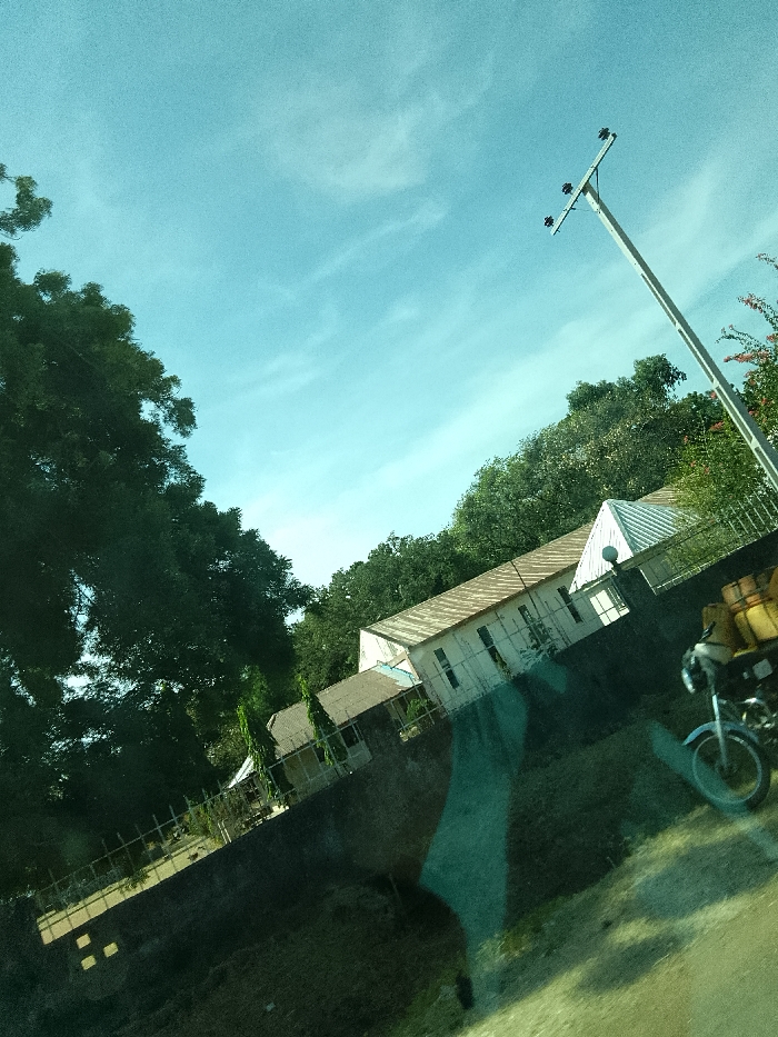 The image depicts an outdoor scene, reportedly in Jibam, Nigeria, under a bright, light blue sky with scattered faint cirrus clouds. The photograph is captured at an angle, exhibiting prominent green and dark reflections across the lower and central areas, consistent with an image taken through a tinted window.
In the midground, a long, single-story building with a light-colored, possibly white, facade and a light-brown corrugated metal roof is visible. The building features multiple rectangular windows. To its left, a smaller, darker-roofed building is partially discernible amidst dense green trees. A portion of a white, pointed roof structure is present to the right of the primary building. A dark, low retaining wall extends along the base of these structures.
On the right side of the frame, a tall, light-colored utility pole with a crossarm supporting four visible insulators stands against the sky. In the lower right foreground, the front section of a light-colored motorcycle is present, displaying a front wheel, fender, and handlebars. Two yellow cylindrical containers, consistent with jerrycans, are affixed to the rear section of the motorcycle, which is mostly out of frame.
Dense green foliage and trees occupy the left side and background of the scene. The immediate foreground consists of an unpaved or dirt surface, partially obscured by the internal reflections. No human subjects are visible in the frame.