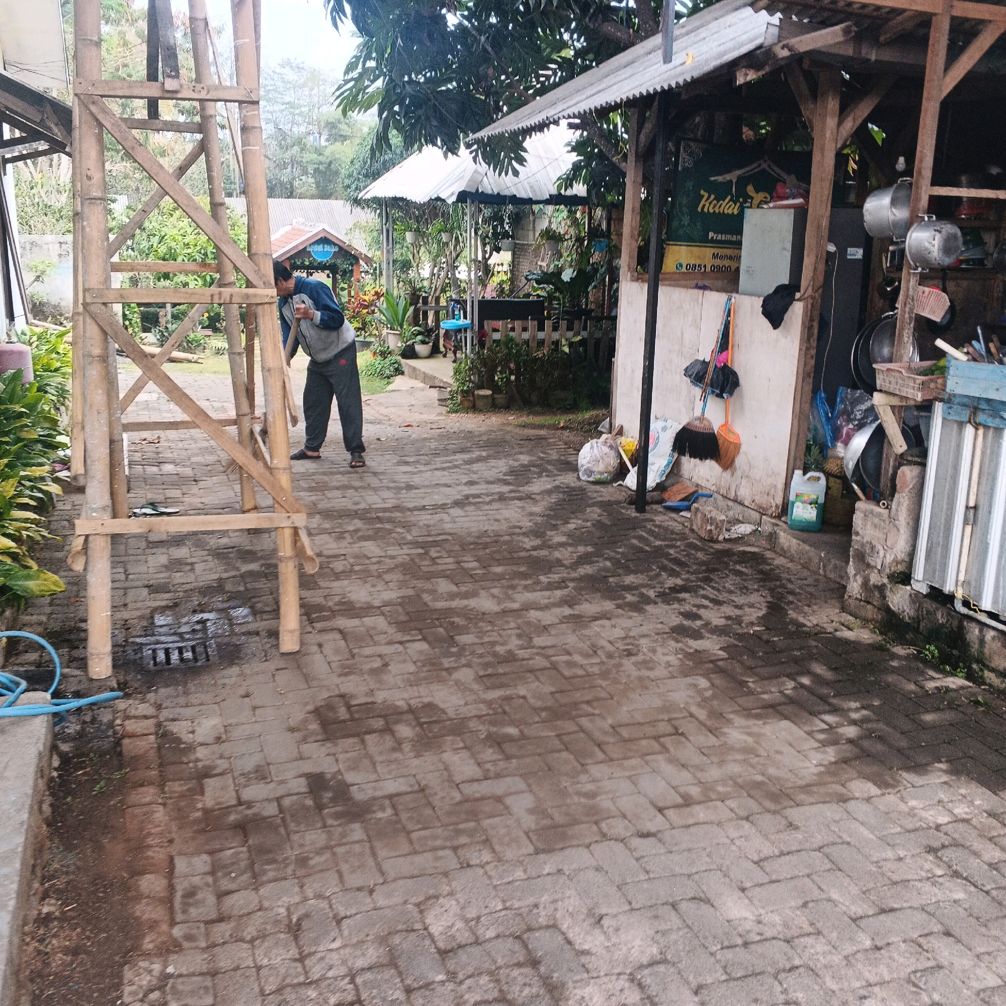 The image shows a person in a blue jacket and gray pants holding a shovel near a wooden structure in Bocek, Indonesia. The person appears to be working, possibly in a garden or outdoor area. The scene is paved with brick pavers, and there are various objects scattered around, including plants, a water hose, and some debris. In the background, there are several open-air structures with thatched roofs, suggesting a market or food stall area. A sign is visible on one of the structures that reads "Kedai". The overall environment appears to be somewhat rustic and outdoorsy.