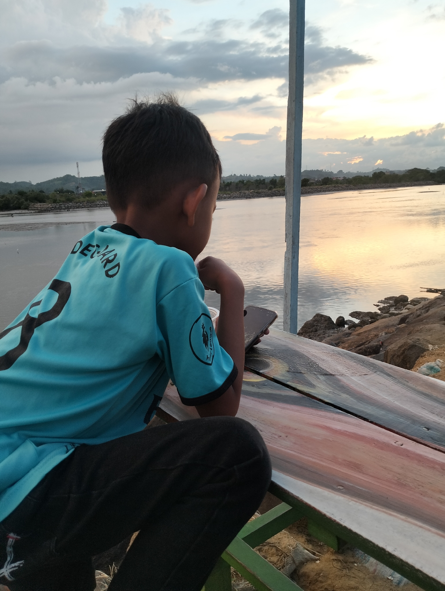 A child sits on a painted bench overlooking a body of water in Lhokseumawe, Indonesia. The child wears a light blue t-shirt with dark sleeves, black pants, and has short dark hair. The t-shirt has the name "DE'NARD" printed on the back and a logo on the sleeve. The bench is painted with multiple colors and made of wood planks. A cell phone sits on the bench next to the child's arm. The water reflects the colors of the sky at sunset. Trees and a structure with a communication tower are visible in the distance. A white pole stands next to the bench. Rocks line the edge of the water.