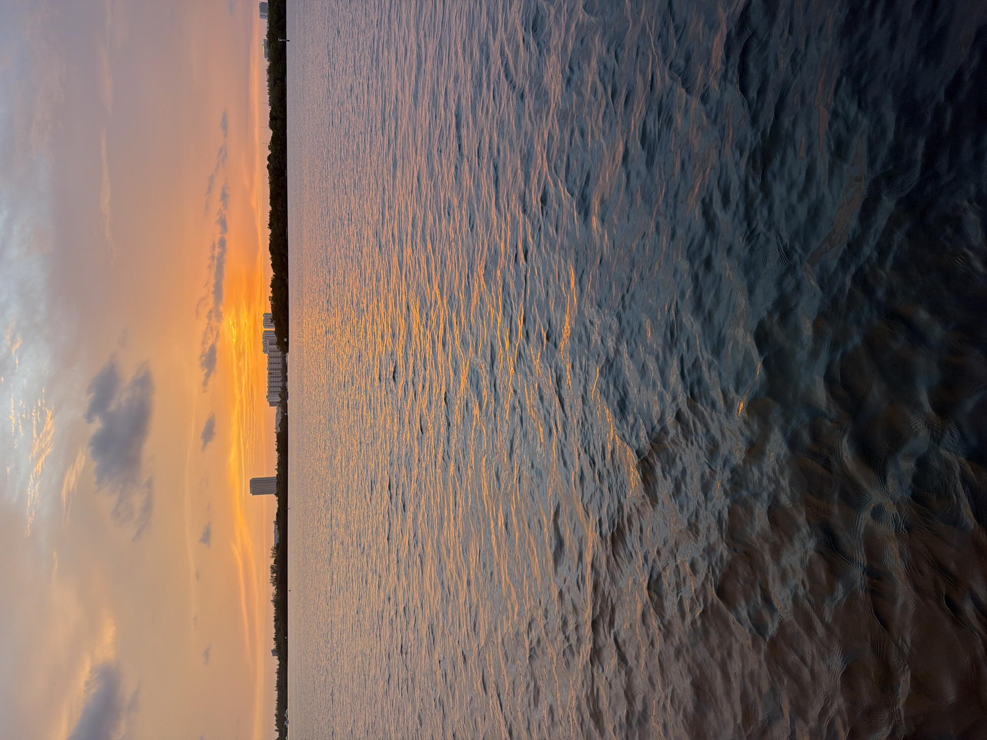 This media file depicts a sunset over a body of water, likely an ocean or large bay, with a city skyline in the distance. The scene is oriented vertically, with the sky occupying the left half of the image and the water the right.
The sky is filled with soft, warm hues of orange, pink, and yellow, indicative of a sunset. Wispy clouds are scattered across the sky, some appearing darker and more defined, while others are lighter and more diffused. A sliver of a dark tree line can be seen separating the sky from the water, with several tall buildings of the city skyline rising above it.
The water exhibits a textured surface with ripples, reflecting the colors of the sunset. The light from the setting sun creates a shimmering golden path across the water, gradually transitioning to darker, more shadowed tones as it moves away from the light source. The overall mood is tranquil and serene, capturing the beauty of a coastal sunset.
The location context provided indicates Miami Beach, United States of America. There is no visible text in the media file. The time of day is clearly sunset. The weather appears calm and clear with scattered clouds.