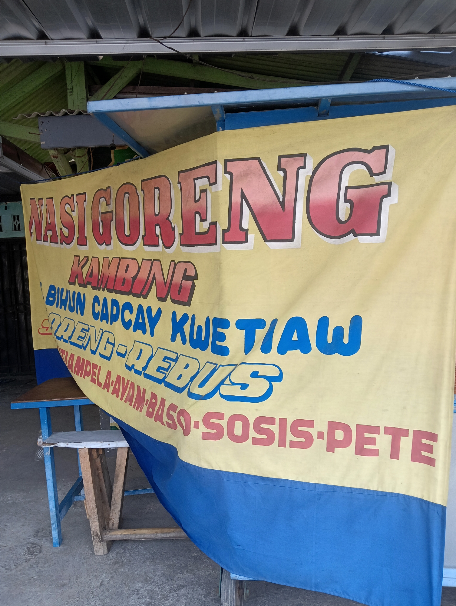 The image shows a yellow banner with blue and red lettering, likely a menu or advertisement for a food stall. The banner displays text such as "NASI GORENG," "KAMBING," "BIHUN CAPCAY KWETIAW," "ORENG-REBUS," "AMPELA AYAM BASO," and "SOSIS-PETE." The banner is hanging in front of a structure with a corrugated metal roof and green wooden supports, typical of an outdoor food vendor in Indonesia. A small blue and brown wooden table and chair are visible in the foreground, suggesting a place for customers to eat. The scene appears to be an outdoor food stall in the city of Ciporang, Indonesia, given the Indonesian language on the banner and the typical setting.
