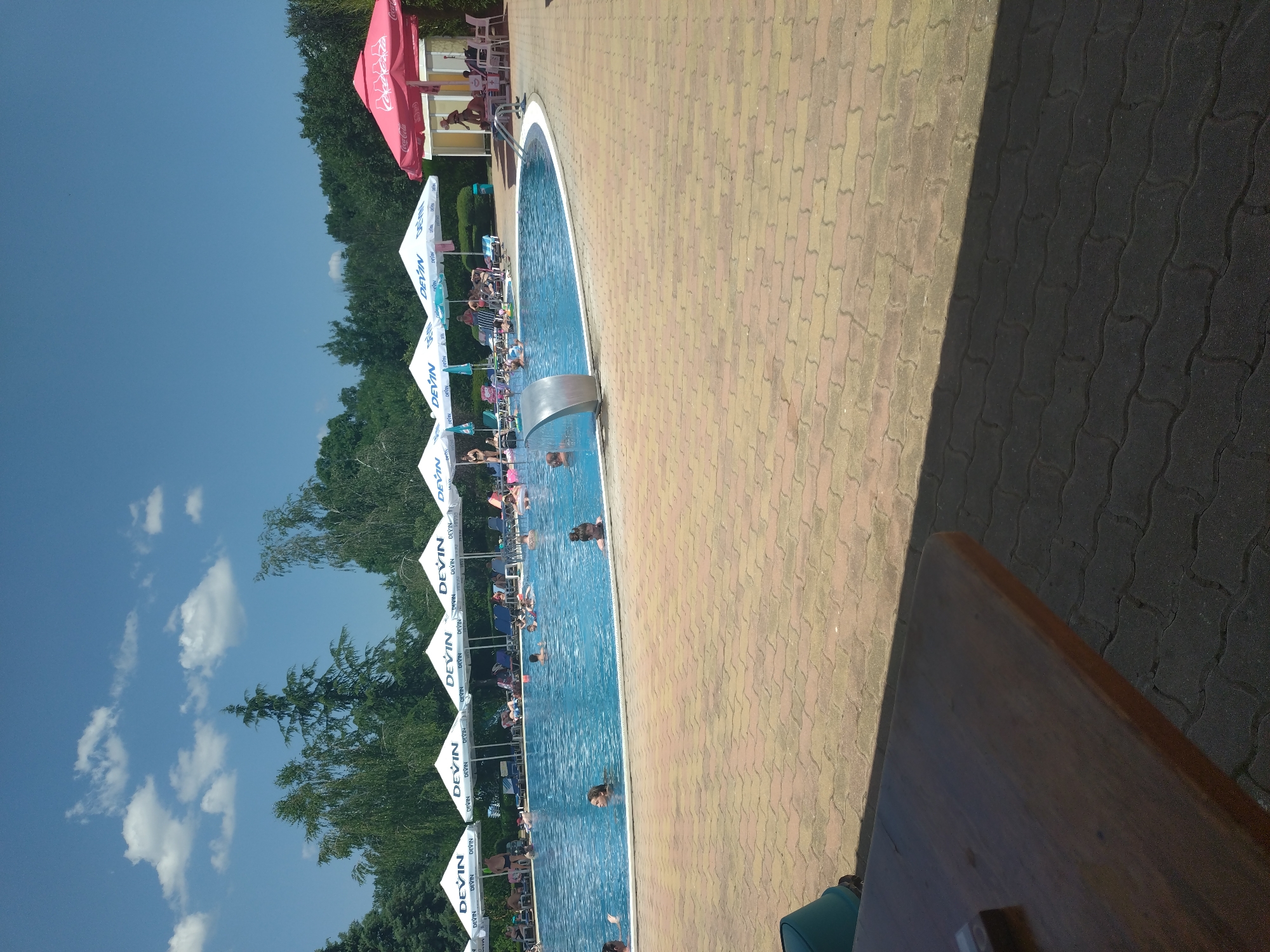 The media file depicts a sunny day at an outdoor swimming pool, likely located in Yagoda, Bulgaria. The scene is vibrant with a clear blue sky dotted with fluffy white clouds. The pool itself is filled with people of various ages enjoying the water. Some are swimming, while others are relaxing or playing. Around the pool, there are lounge chairs and umbrellas, some of which are branded with "DEVIN" and "Coca-Cola," suggesting a commercial establishment like a water park or resort. The poolside is paved with interlocking bricks in shades of yellow and brown. To the right of the pool, a dark shadow suggests the presence of an overhang or structure, and a wooden table occupies the foreground, implying an area for dining or relaxation. The overall atmosphere appears cheerful and relaxed, indicative of a leisure activity on a warm day.