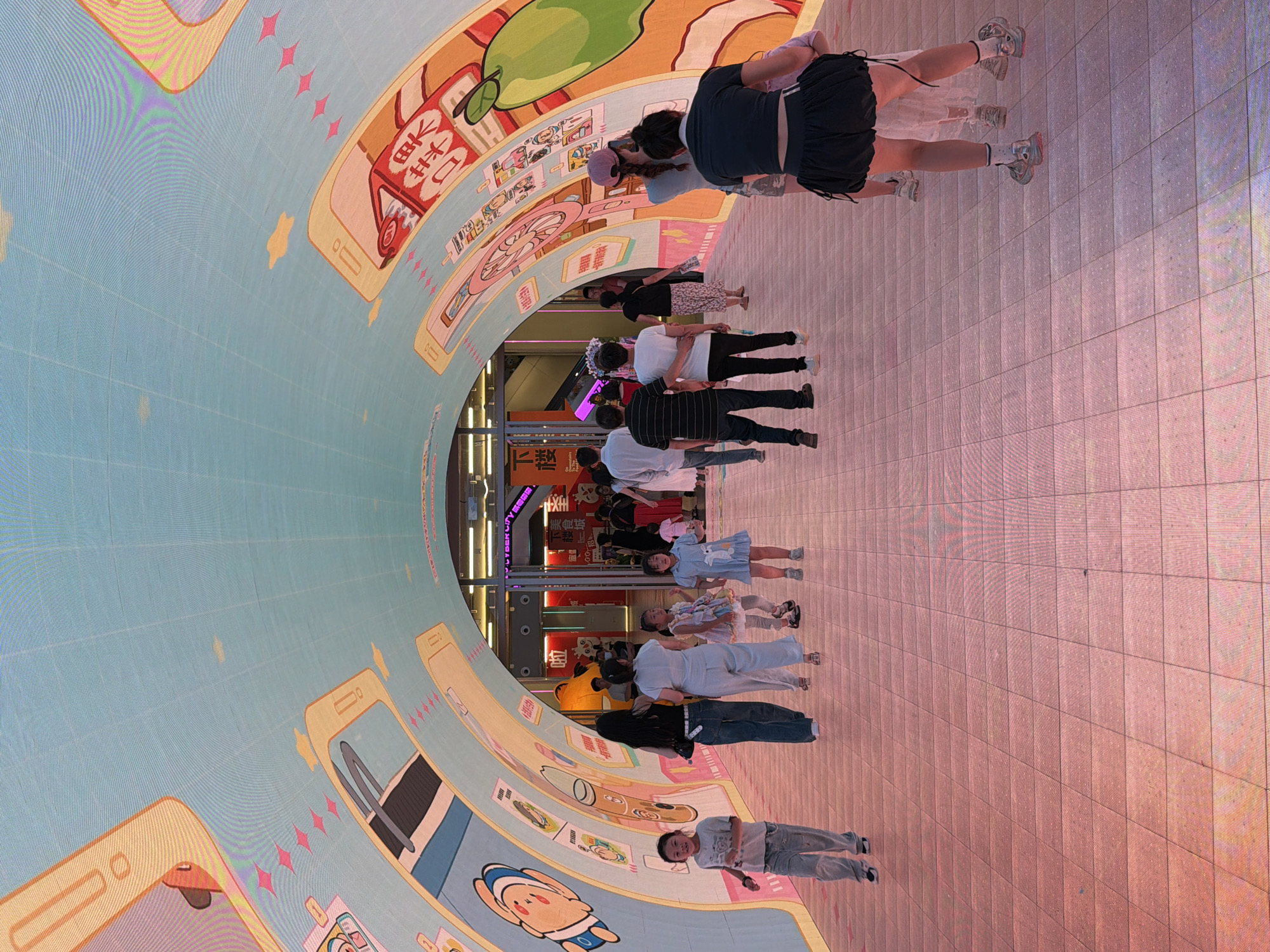 The image captures a group of people, including adults and children, standing on a brightly lit floor. The scene appears to be an indoor digital art installation or a large-scale projection, given the curved, illuminated surface displaying cartoonish graphics and text. The graphics feature stylized representations of mobile phones, stars, and other whimsical elements. The people are positioned in a line or a small group, facing towards the curved screen, suggesting they are observing or interacting with the display.
Some individuals are looking directly at the screen, while others might be posing for a photo. The mood seems casual and lighthearted, as indicated by the presence of children and the overall vibrant and colorful environment. The lighting is artificial and strong, casting a soft pinkish hue on the floor. The time of day is likely evening or night, or an indoor setting with controlled lighting. The presence of Chinese text on signs in the background, particularly "CYBERCITY" and some characters that appear to be "幸福", suggests the location is in China, possibly in a commercial or entertainment district within Yuzhong District. The overall impression is a modern, engaging space designed for public enjoyment and visual stimulation.