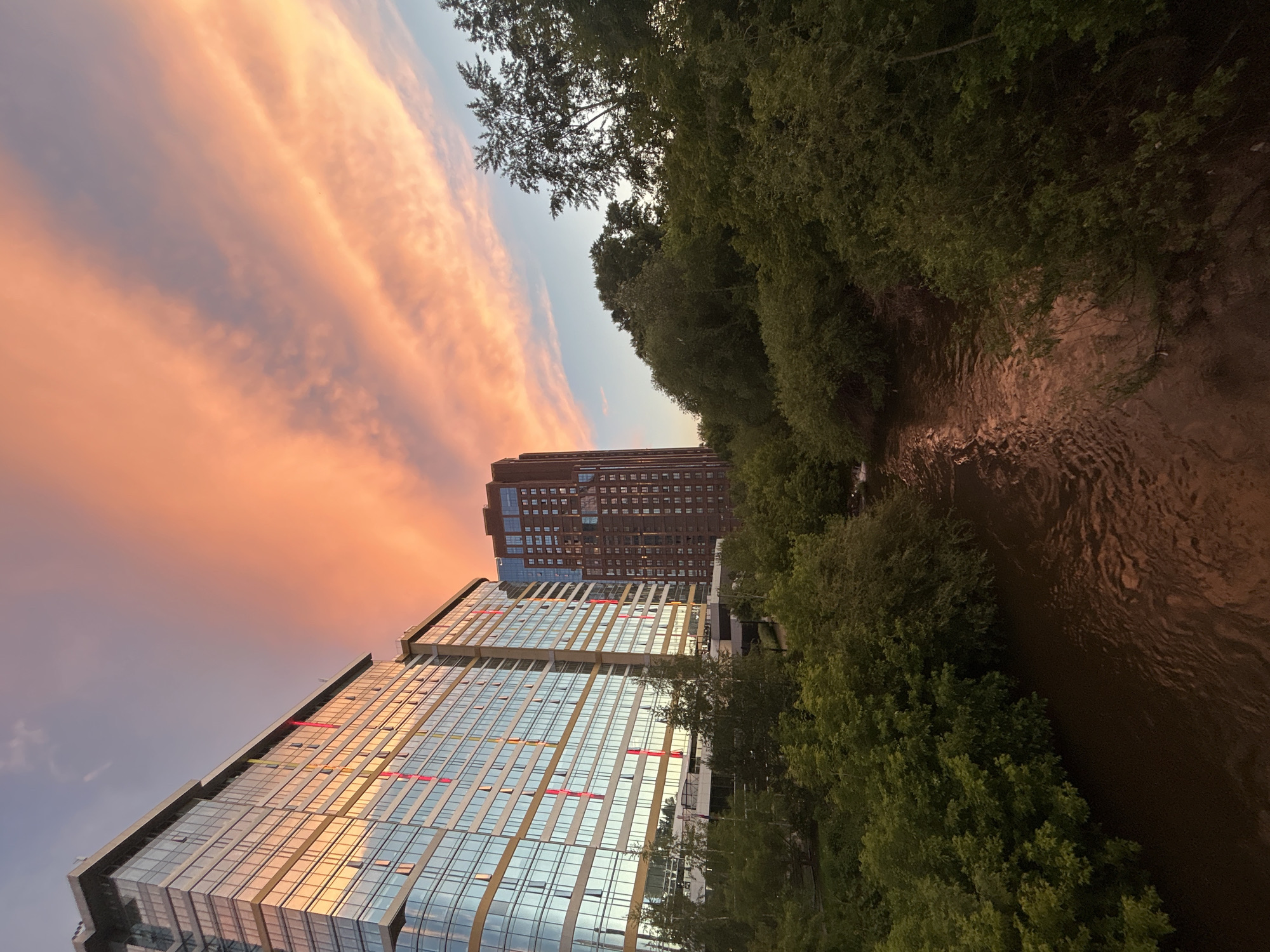This image depicts an urban landscape in Yekaterinburg, Russia, at twilight, characterized by an intensely colored sky, modern and traditional high-rise buildings, dense vegetation, and a river. The specific location is along the Iset River, featuring the "Republic" residential complex prominently on the left.
The sky occupies the upper portion, displaying striated altocumulus clouds illuminated by the setting sun, creating vibrant orange and pink hues that transition to a pale blue-grey gradient towards the upper right.
On the left, a multi-story, contemporary residential building, identified as part of the "Republic" complex, features a facade primarily composed of reflective glass panels. These panels mirror the warm light of the sky in golden-orange tones and also reflect cooler blue light on lower sections. Vertical red and yellow decorative elements are intermittently visible on the building's exterior. To the right and slightly behind, a taller, slender skyscraper constructed from dark brown brick or concrete displays a uniform grid of numerous small, rectangular windows.
A dense band of lush, green deciduous trees and shrubs forms a natural barrier across the mid-ground, obscuring the base of the buildings and framing the water body below. The foliage appears thick and healthy.
A moderately wide, brown-colored river, identified as the Iset River, flows through the lower-center of the frame, partially visible between the vegetation. Its surface exhibits gentle ripples, with subtle reflections of the twilight sky in muted orange and brown tones.
No individuals, animals, or vehicles are present or discernible within the scene. The perspective suggests the image was captured from an elevated position, likely a bridge or a higher vantage point overlooking the river and the urban development.