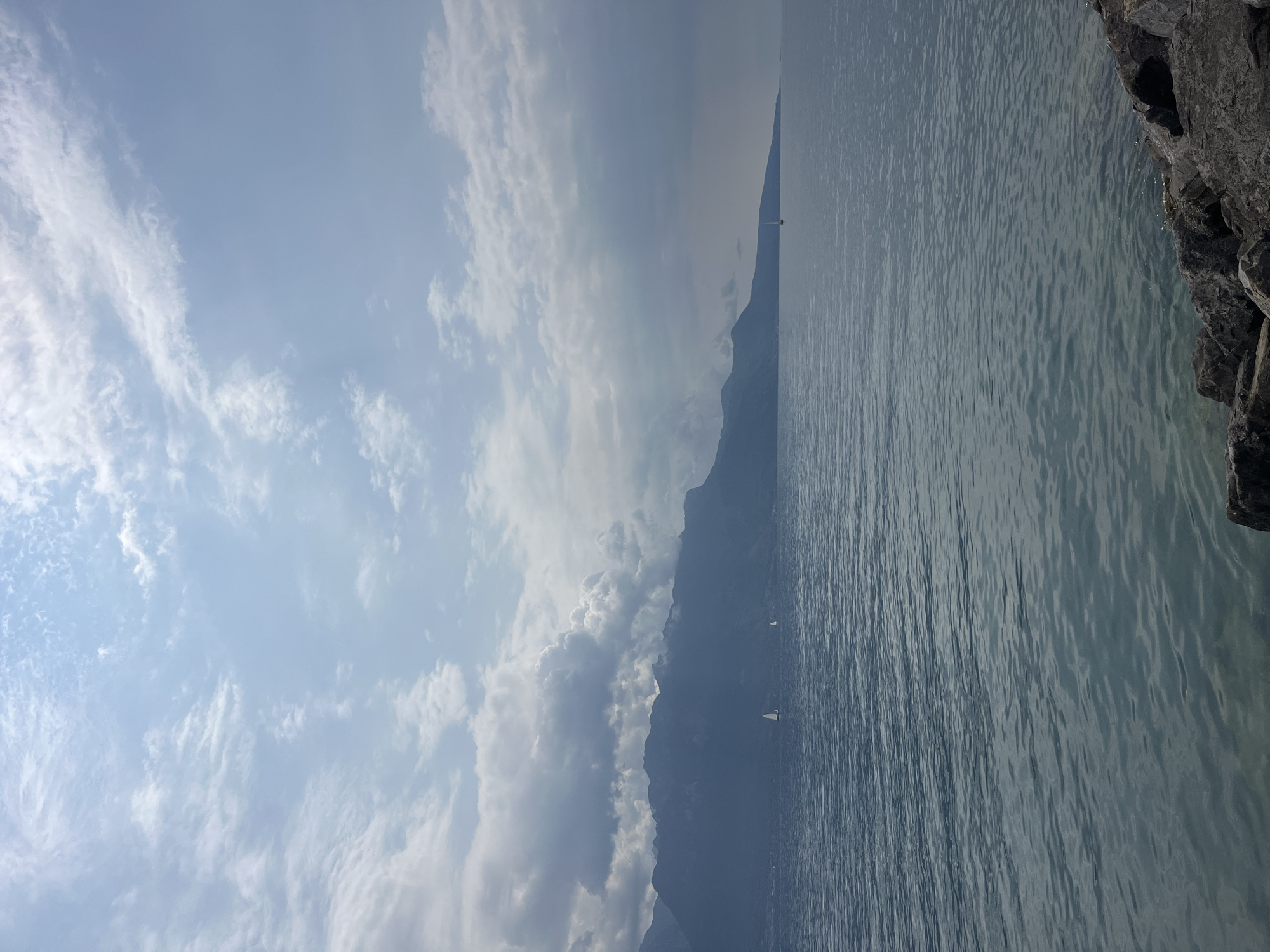 The image depicts a wide shot of a large body of water, likely Lake Geneva, with a mountainous landscape in the background. The sky is partly cloudy, with sunlight filtering through in places, creating a dynamic interplay of light and shadow. In the middle distance, two sailboats are visible on the water, adding a sense of scale and activity to the scene. The foreground features a rocky shoreline, composed of large, dark gray stones. This view is characteristic of the lakeside promenade in Vevey, Switzerland, known for its stunning views of the lake and the surrounding Alps. No people are visible in the frame. The overall impression is one of serene natural beauty, with the rugged mountains contrasting with the calm water.