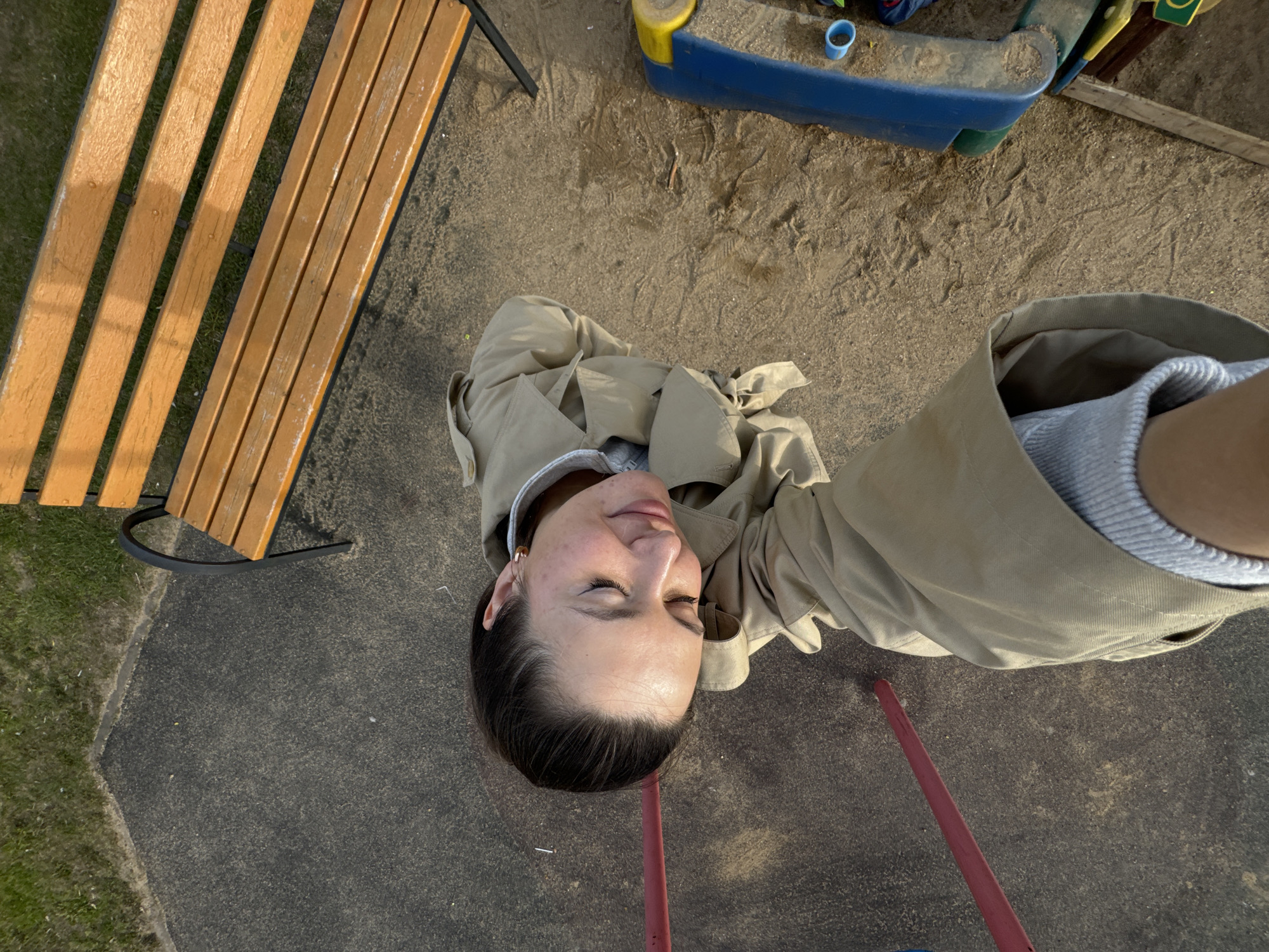 The image, taken from an overhead perspective, shows a woman in a tan trench coat standing in what appears to be a children's playground. A wooden bench is visible on the left, and part of a playground structure, possibly a sandbox, is in the upper right corner. The ground is a mix of pavement and sand. The woman's face is centered in the frame; she has her eyes closed. The location may be Akulovo (Gp Kubinka), Russia, based on provided metadata.