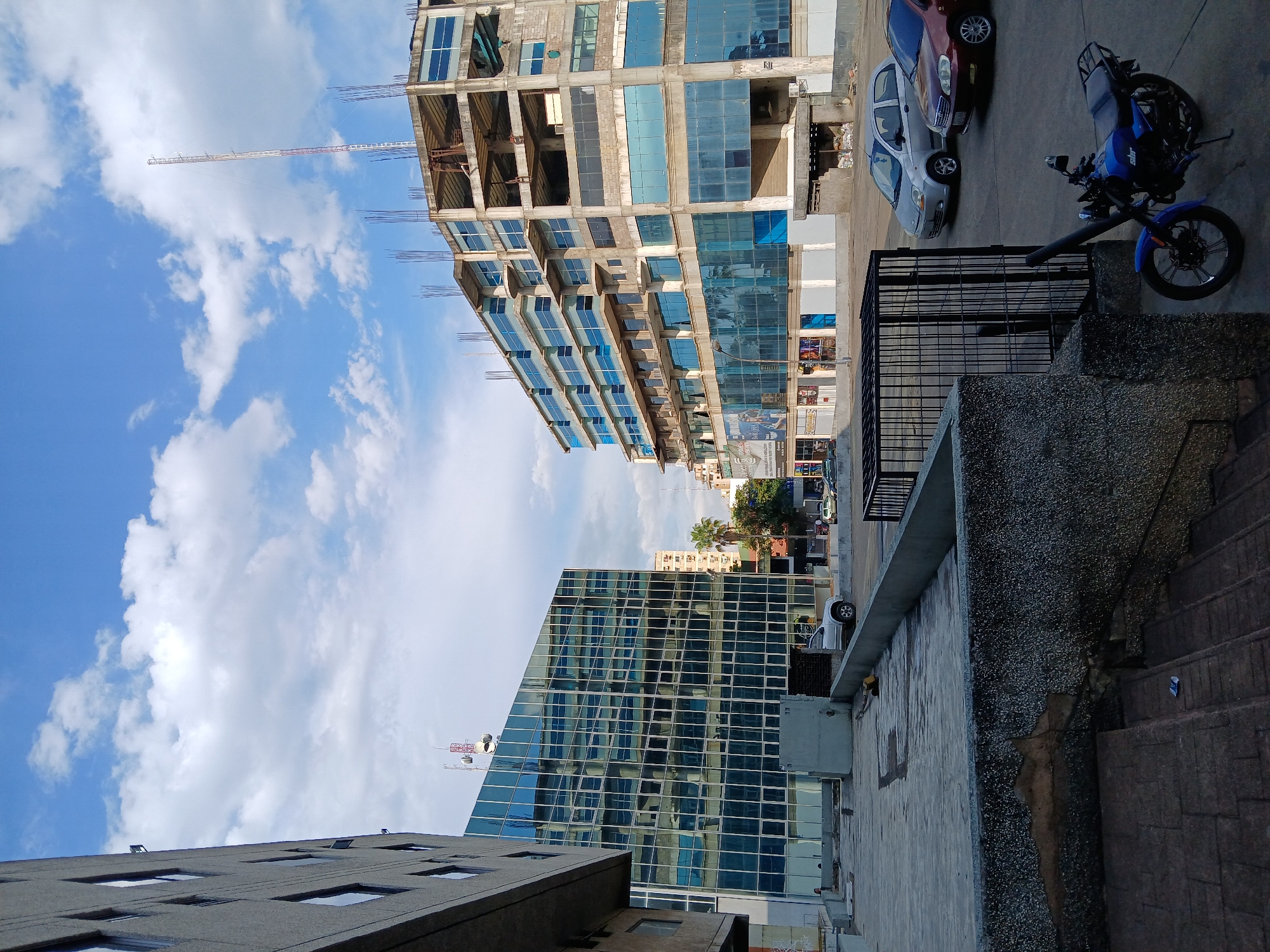 This urban landscape photograph is rotated 90 degrees clockwise, presenting a street scene dominated by buildings and vehicles under a bright sky.
On the left side of the frame, a clear blue sky with scattered white cumulus clouds fills much of the view. A tall, white and red construction crane extends diagonally upwards from behind a building into this sky.
In the upper-right portion, a multi-story building is under active construction. Its raw concrete structural framework is visible, with rebar protruding from unfinished sections. Several floors are partially clad with modern blue reflective glass windows, suggesting its future appearance. At its base, at street level, a large sign is visible with text including "GUYANA MALL" and "CANAIMA", along with other unreadable details, indicating a commercial development.
In the lower-left, another multi-story building stands completed, featuring a contemporary design with a facade entirely composed of blue reflective glass panels. This building reflects the sky and surrounding environment. A small communication tower or antenna is visible on its roof.
The right side of the image depicts street level, with a paved area for parking. A prominent blue motorcycle is parked in the immediate foreground, next to a black metal fence. Behind it, a light-colored sedan and a maroon sedan are also parked. The immediate foreground on the far right consists of rough, textured concrete structures, possibly a wall or steps.
The bright lighting and clear sky suggest it is daytime with fair weather. No people are visible in the scene, which captures a moment of an active urban development in Ciudad Guayana, Venezuela.