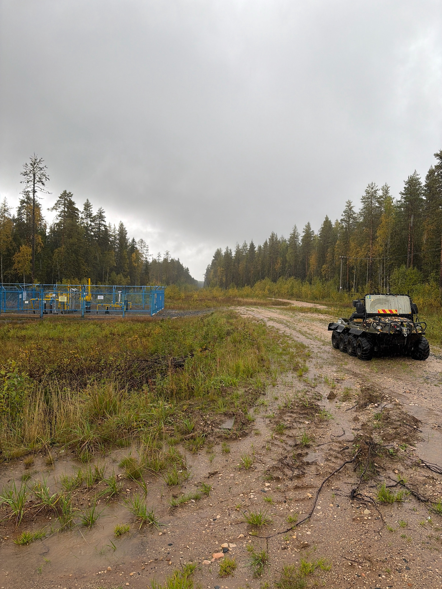 The image depicts an outdoor scene on an overcast day in Yemtsa, Russia, likely near an industrial site. In the foreground, a wet, gravelly track shows tire marks and scattered debris, with puddles of water reflecting the sky. To the left, a fenced-off area contains industrial equipment, characterized by yellow pipes and machinery enclosed within a blue metal fence. This facility is situated amidst tall grasses and some autumn foliage. In the mid-ground and extending into the background, a dense forest of pine and birch trees lines the path, with the trees displaying a mix of green and yellowing leaves, indicative of the autumn season. To the right, a multi-wheeled amphibious vehicle, camouflaged and bearing a Swedish flag and yellow and red reflective tape, is parked on the track. The vehicle appears to be equipped for off-road or rugged terrain. The overall atmosphere is one of a remote, industrial area within a natural, wooded environment, under a cloudy sky.
