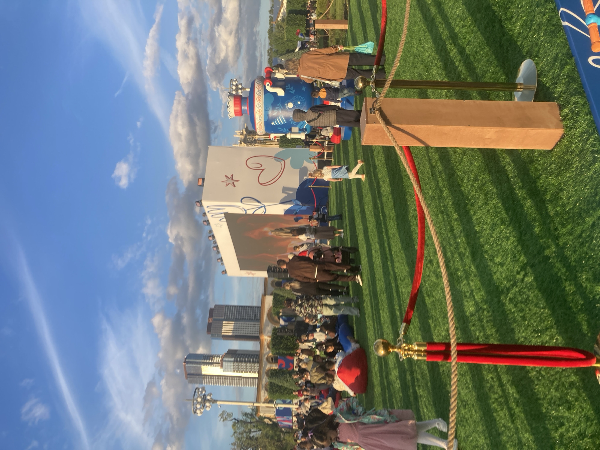An outdoor public event unfolds on a large expanse of artificial green turf under a clear blue sky dotted with scattered white clouds. The time appears to be late afternoon or early evening, characterized by the golden sunlight and long shadows cast across the grass.
Numerous people, including adults and children, are gathered, some standing and many seated on the grass or blankets. Their attire is casual. The focal point of the gathering appears to be a large white screen or billboard, positioned centrally. This screen features a prominent red heart and a red star, along with partially visible stylized text and what seems to be a character or scene.
To the right of the screen stands a large, blue, intricately decorated object, possibly a stylized robot or figure, adorned with red and white accents. In the immediate foreground, a red velvet rope barrier with gold stanchions and a separate natural fiber rope demarcate an area, along with a wooden block-like barrier. On the far right, a blue surfboard-like object with partially visible white text that reads "My Little..." lies on the grass.
In the background, a modern urban skyline with several high-rise buildings is visible, along with streetlights. The overall atmosphere is relaxed, suggesting a communal, family-friendly event or festival in a city setting. The event is taking place in Moscow, Russia.