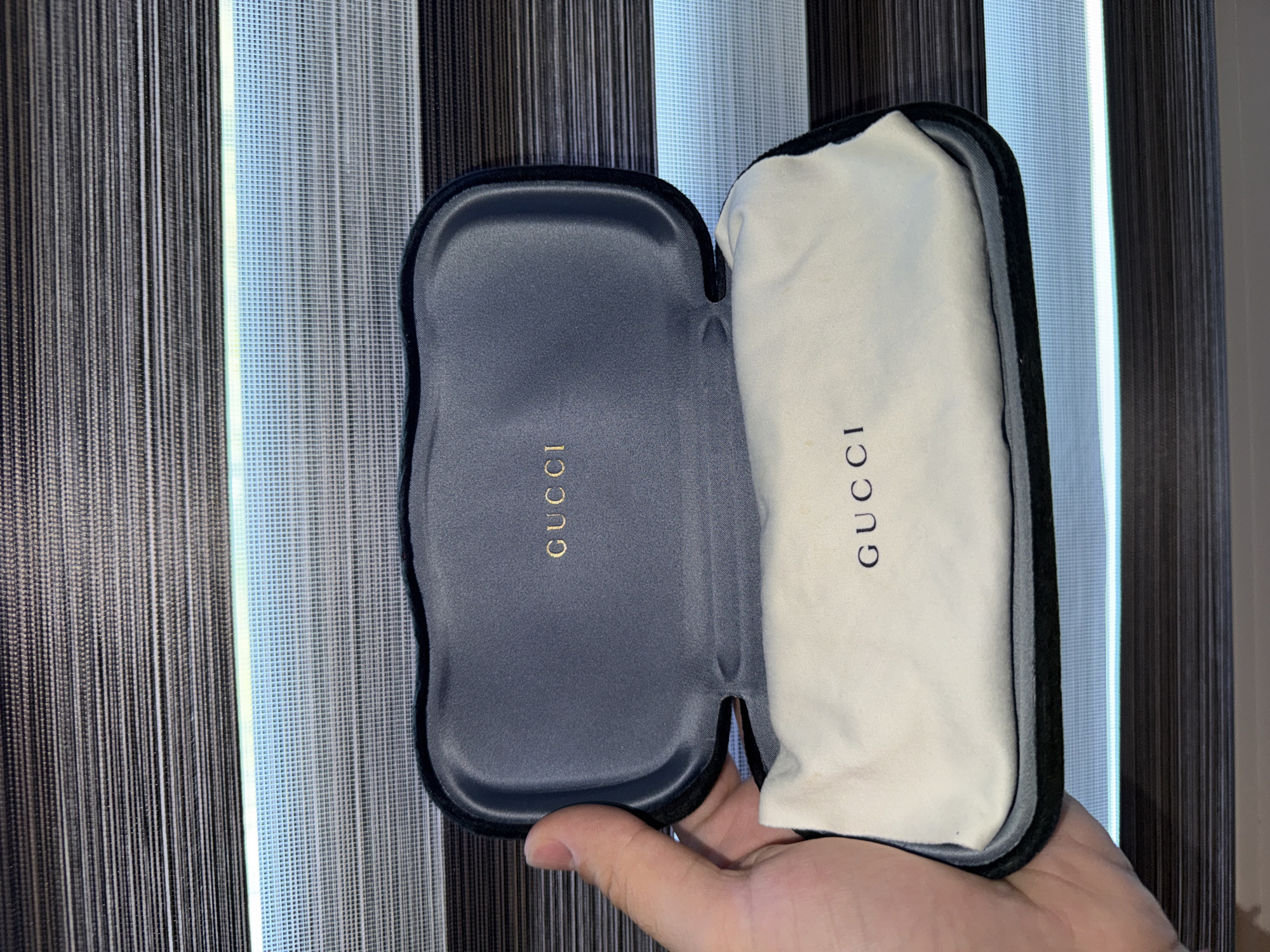 A person's hand holds open a dark gray velvet-lined Gucci glasses case. The case contains a white cloth bag with the Gucci logo printed on it. The background is a set of horizontal blinds with varying shades of gray and light blue. The scene appears to be indoors, possibly in a home environment in Varna, Bulgaria, though this location cannot be confirmed from the image alone.