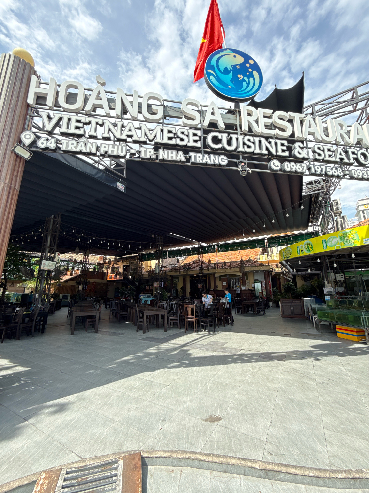 This is a photograph of the entrance to Hoang Sa Restaurant, a Vietnamese cuisine and seafood establishment located at 64 Tran Phu Street in Nha Trang, Vietnam. A large, white, illuminated sign above the entrance reads "HOANG SA VIETNAMESE CUISINE & SEAFOOD" along with a phone number. A Vietnamese flag and a circular logo featuring a blue fish are also visible on the sign. The restaurant features an outdoor seating area with numerous dark wooden tables and chairs, covered by a large black awning. The floor is paved with light-colored tiles, and there are overhead lights strung across the dining area. In the background, the architecture of the building appears to be traditional Vietnamese style, with tiled roofs. A few people are visible within the restaurant and near the entrance, some appearing to be staff and others customers. The environment suggests a sunny day with some shadows cast by the overhead covering and trees.