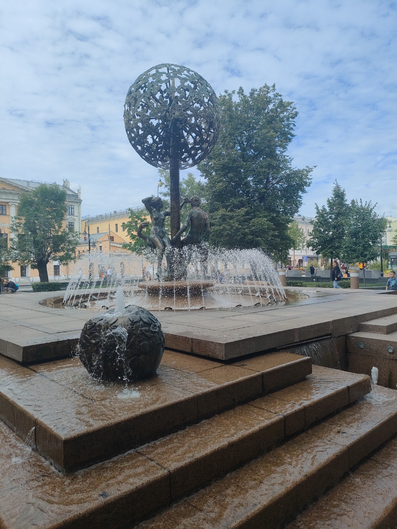 The image depicts a fountain in a city square, likely in an urban park or plaza. The central feature is a large, ornate fountain with sculpted figures and a spherical, openwork element at the top, resembling a globe adorned with leaves or abstract shapes. Water cascades from multiple tiers of the fountain, creating a dynamic and refreshing visual. In the foreground, a smaller spherical element, also with water flowing over it, sits on a tiered stone structure, suggesting a multi-level water feature. The surrounding environment includes paved walkways, stone steps leading down from the fountain, and lush green trees providing shade and a natural backdrop. Buildings with traditional architecture, featuring light-colored facades and arched windows, are visible in the background, indicating a presence of urban development. Several people are scattered throughout the scene, some sitting on benches, others walking, and children are present, suggesting a public and recreational space. The sky is partly cloudy, with a mix of blue sky and white clouds, indicating moderate weather conditions. The overall atmosphere is one of urban serenity and public enjoyment.