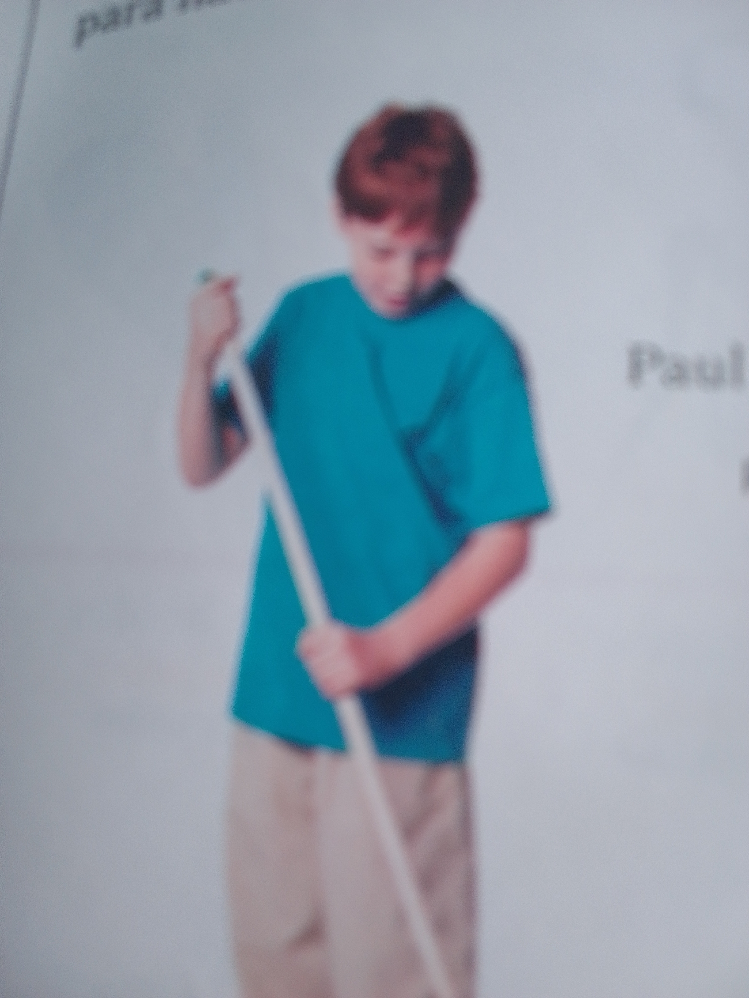 A young boy with reddish-brown hair, wearing a teal t-shirt and tan pants, stands holding a white stick. The boy is looking downwards. Text on the background reads "Paul". The image is blurry and appears to be a photograph taken indoors with a white background. The location of La Tagua, Venezuela is not depicted or suggested in the image.