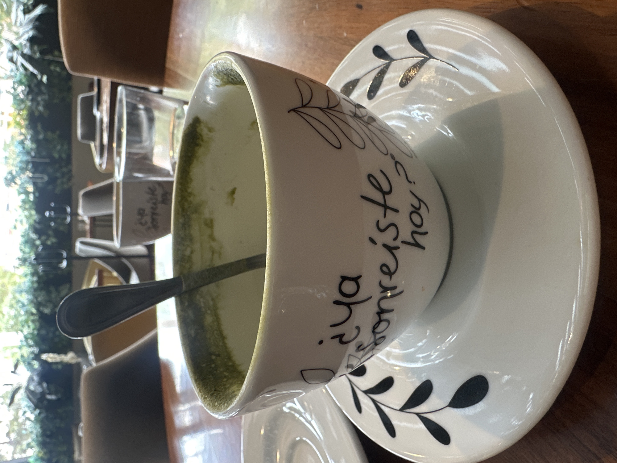 A slightly angled close-up shot presents a coffee cup and saucer on a wooden table in what appears to be a cafe setting. The cup, primarily white with hand-drawn black leaf designs, contains remnants of a greenish beverage. A silver spoon rests inside the cup. The saucer is white with matching black leaf designs. On the cup, there is text in Spanish, "¿ya sonreíste hoy?" which translates to "Have you smiled today?" A partial view of other cups and saucers suggests additional table settings. The scene is located in Huixquilucan de Degollado, Mexico. The environment appears bright, possibly from natural light coming through a window, as foliage is seen in the blurry background on the left.