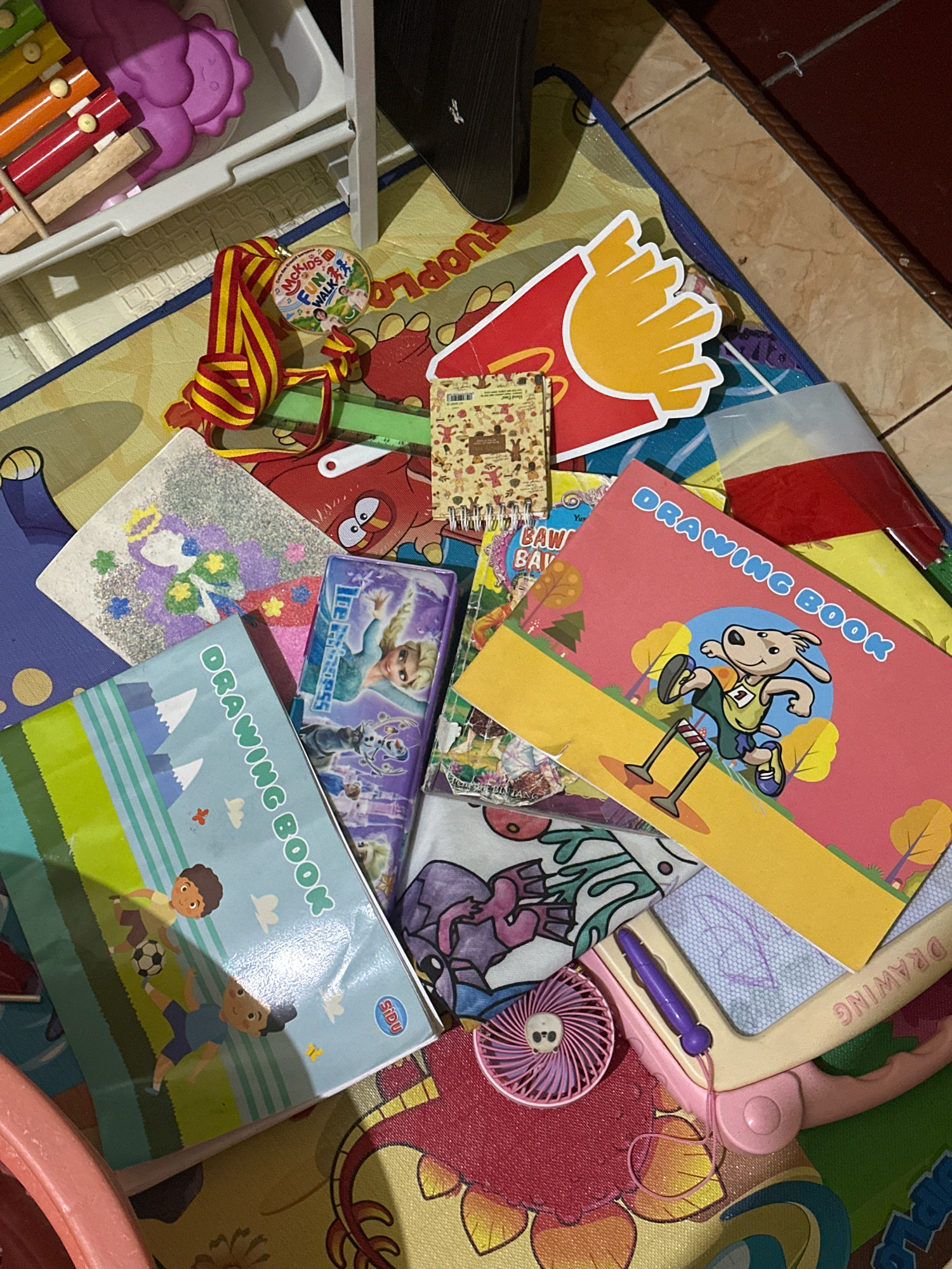 This image depicts a collection of children's toys and school supplies scattered on a colorful mat, likely on the floor of a room in Ngasem, Indonesia. The objects include a purple plastic xylophone with colored bars, a McDonald's Happy Meal-themed drawing pad with french fries graphics, a medal with "McKids Fun Walk" text, two drawing books with cartoon characters and landscapes, a small purple electric fan, a pink drawing tablet with a stylus, and various other colorful papers and toy items. The lighting suggests an indoor environment, possibly a playroom or a child's bedroom.