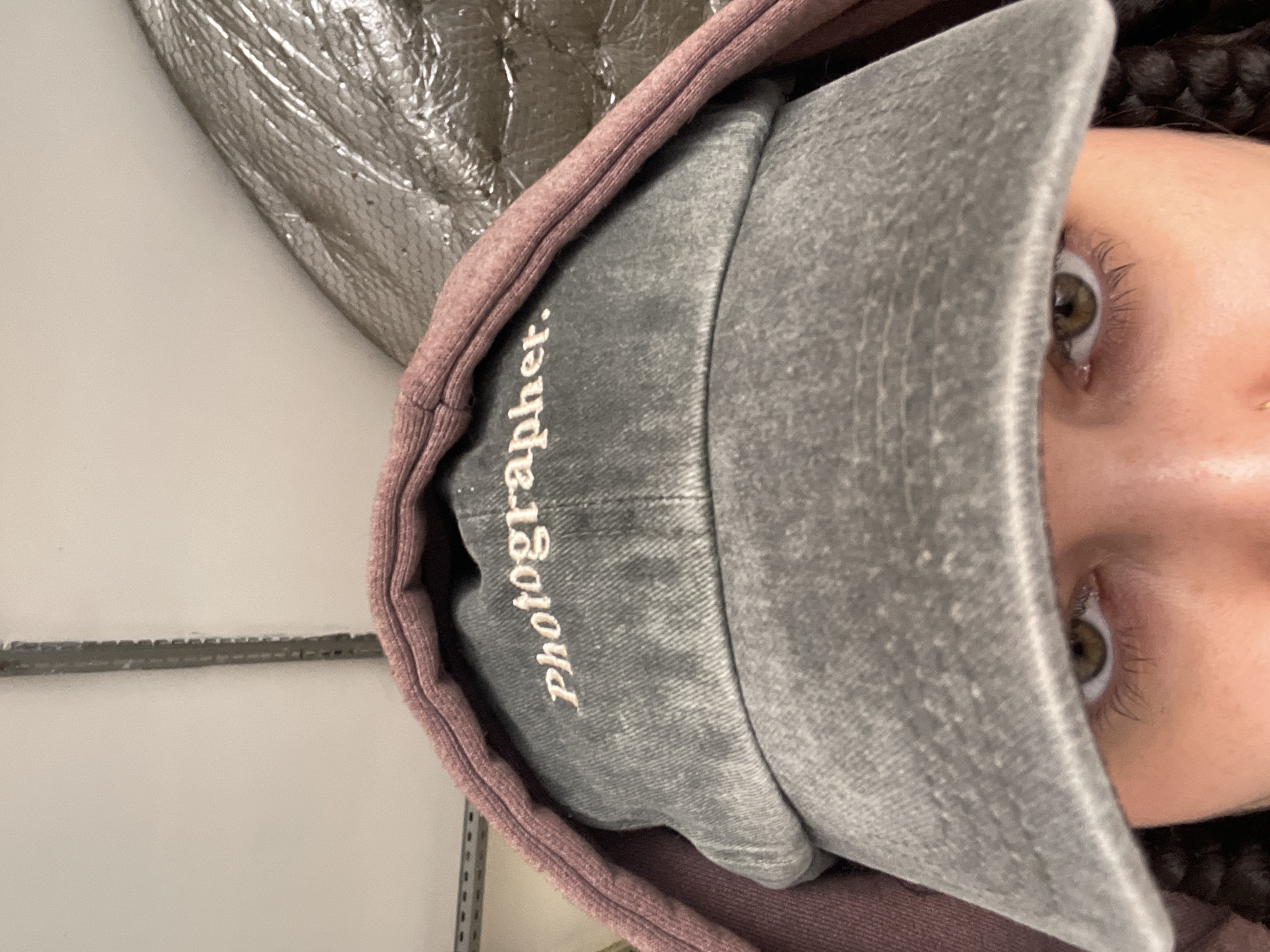 A close-up image of a person wearing a grey cap with "Photographer." embroidered on the front, and a mauve hoodie, is visible. The person has light skin, with brown eyes, and dark hair is visible above the hoodie. Part of a metal shelving unit and a quilted metallic object are in the background. The location in Moscow, Russia, is not discernible from the image.