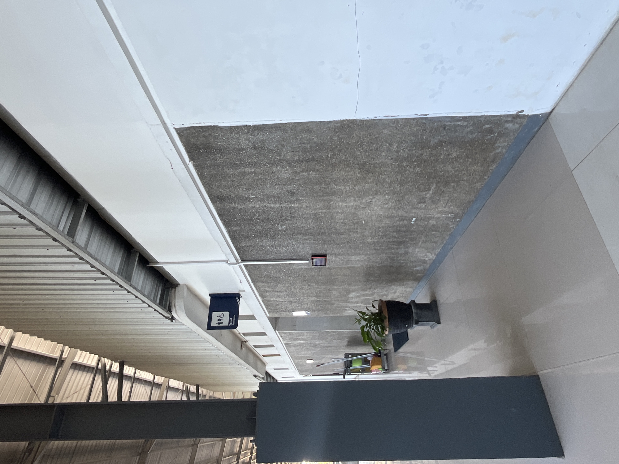 An outdoor hallway with a corrugated metal roof extends to the right, supported by a dark grey pillar on the left. The hallway floor is tiled in a light beige color. The walls on the right are a rough, grey texture with some white paint. A dark blue sign indicating "Toilet" and featuring figures representing men, women, and a person in a wheelchair is mounted on the white wall. Further down the hallway, a cleaning cart with a yellow bucket and mop is visible. A potted plant sits on a pedestal near the cleaning cart. A red fire alarm box is mounted on the grey wall. The scene appears to be in a public area, possibly a transportation hub or a commercial building in Desa Tambakreja, Indonesia, given the architectural style and the tropical foliage.