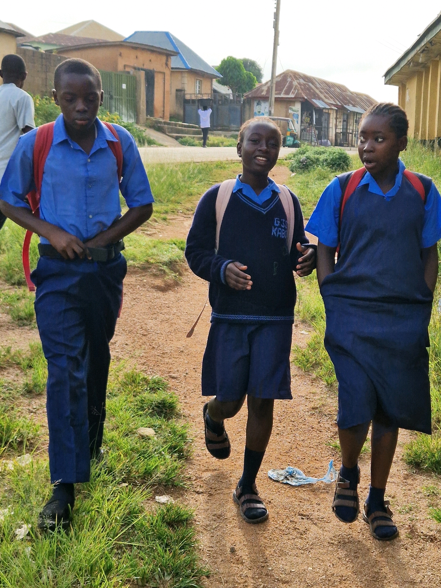 Three children in school uniforms walk along a dirt path lined with dry grass. The child on the left, a boy wearing a blue short-sleeved shirt, dark blue pants, and a red backpack, is adjusting his black belt. The child in the middle, a girl in a navy blue v-neck sweater over a blue shirt and dark blue shorts, smiles. The child on the right, a girl in a blue short-sleeved shirt with a navy blue vest and dark blue skirt, looks off to the side with her mouth slightly open. In the background, a few buildings with corrugated metal roofs are visible, suggesting a semi-urban or rural setting within the city of Kabong, Nigeria. The overall lighting indicates it is daytime.