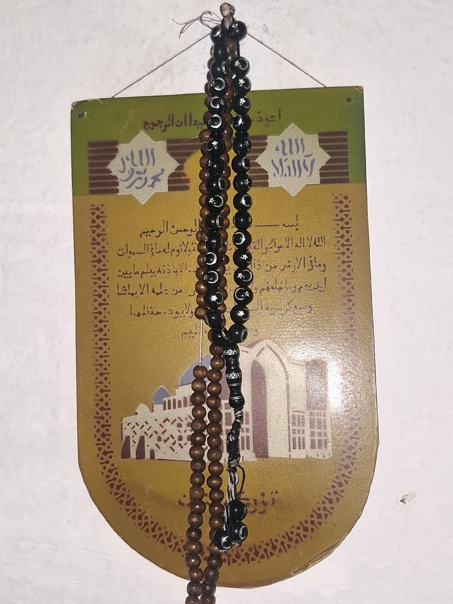 The image shows a close-up of Islamic prayer beads (tasbih) hanging against a decorative plaque. The plaque has Arabic calligraphy and a stylized image of a mosque. The prayer beads are of two types: one set is made of dark, possibly black, beads with carved crescent moon and star symbols, and another set is made of smaller, light brown wooden beads. The background appears to be a white wall. The image does not contain any information that would indicate its location in Almaty, Kazakhstan, or any other specific city. There are no people, actions, or interactions depicted.