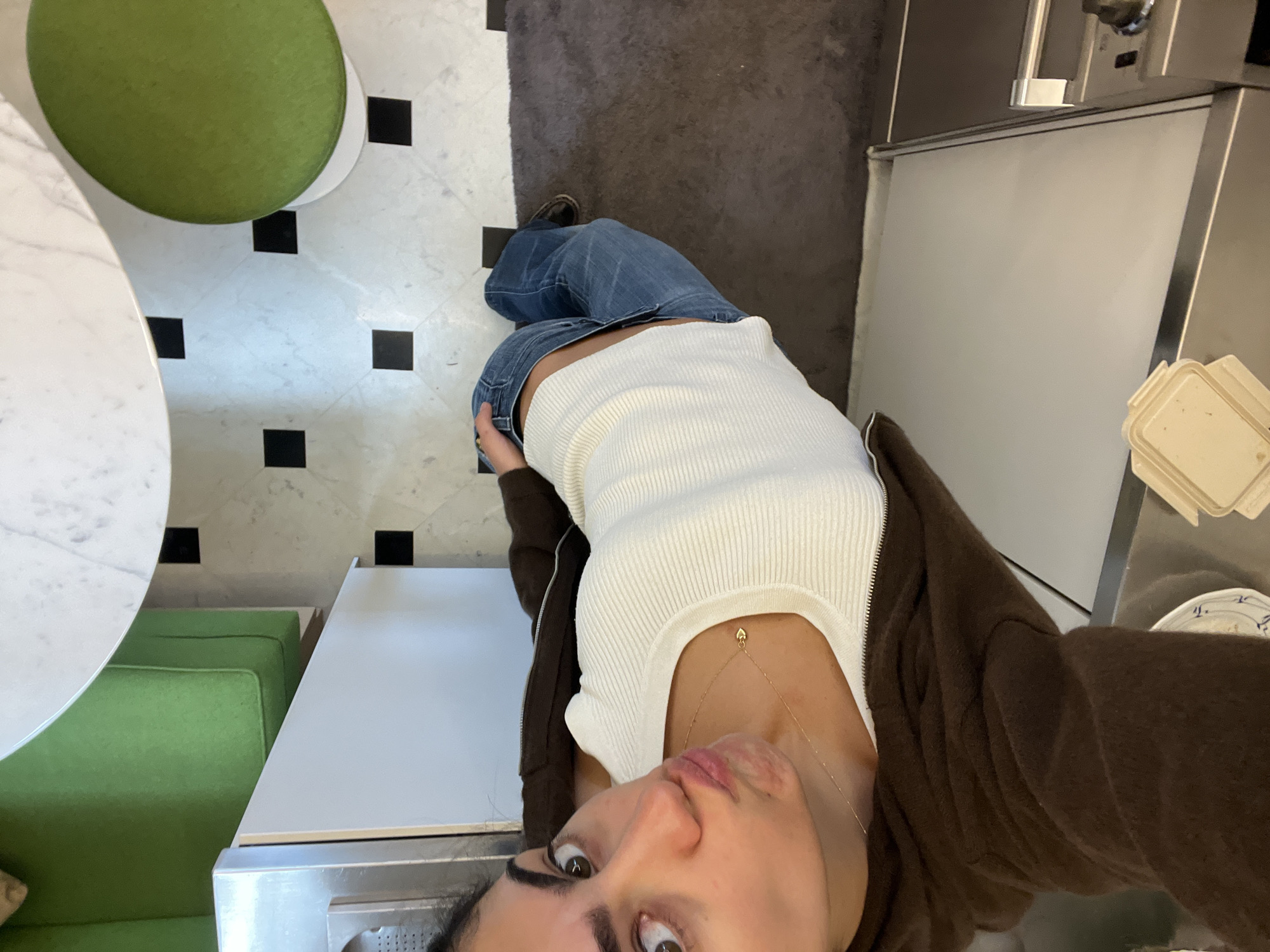 The image is a high-angle selfie taken indoors, looking down at a person and their immediate surroundings. The subject, appearing to be female, has dark hair and is looking directly up at the camera with a neutral expression. She is wearing a white ribbed tank top, light blue denim jeans, and a dark brown zip-up hoodie worn open. A delicate gold chain with a small heart charm is visible around her neck.
The setting appears to be a cafe or restaurant in Paris, France. The flooring transitions from a white tiled area with black square inserts to a dark grey or black textured floor. To the left, there is a round white marble-top table and vibrant green upholstered booth seating. On the right, a light-colored counter, possibly stainless steel, is visible, with beige takeout containers and a white plate with blue patterns placed on or nearby. In the background, commercial kitchen or service equipment can be seen, possibly including a refrigerator unit.
The person is posed for the photo, with no other specific activity evident. The lighting suggests an indoor setting, likely during the day. No legible text is visible within the frame.