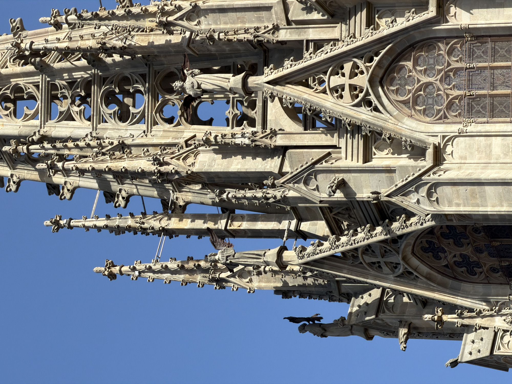 The image shows a close-up, low-angle view of the spires and Gothic architecture of the Barcelona Cathedral, located in the Gothic Quarter of Barcelona, Spain. The sky is a clear, vibrant blue. The stone facade is intricately carved with pointed arches, gargoyles, and statues. Two statues of angels are visible on the spires. The sunlight casts sharp shadows, highlighting the detailed stonework and the vertical lines of the building. The architectural style is characteristic of Gothic cathedrals, with elaborate ornamentation and pointed spires reaching towards the sky.