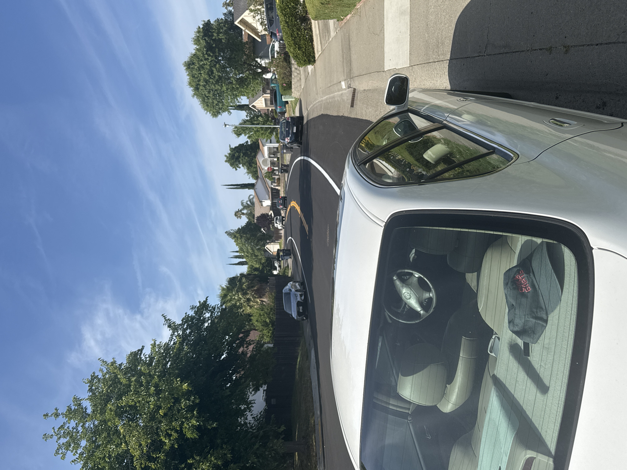 The image shows a residential street in Citrus Heights, USA, on a bright, sunny day. A white car, from which the photo appears to be taken, is prominently featured on the right side of the frame. Its interior is visible through the windshield, revealing cream-colored seats, a steering wheel, and a black hat with red text on the front passenger seat.
The street ahead is newly paved with dark asphalt, clearly marked with a continuous white line and a yellow line on the left side of the lane. A concrete sidewalk runs along the right side of the road. On both sides of the street, houses are nestled among lush green trees and hedges, and several other vehicles are visible further in the distance. The sky is a vibrant blue with sparse, wispy white clouds, indicating clear weather. The strong sunlight casting distinct shadows suggests it is late morning or early afternoon, depicting a typical suburban environment. No people are visible.