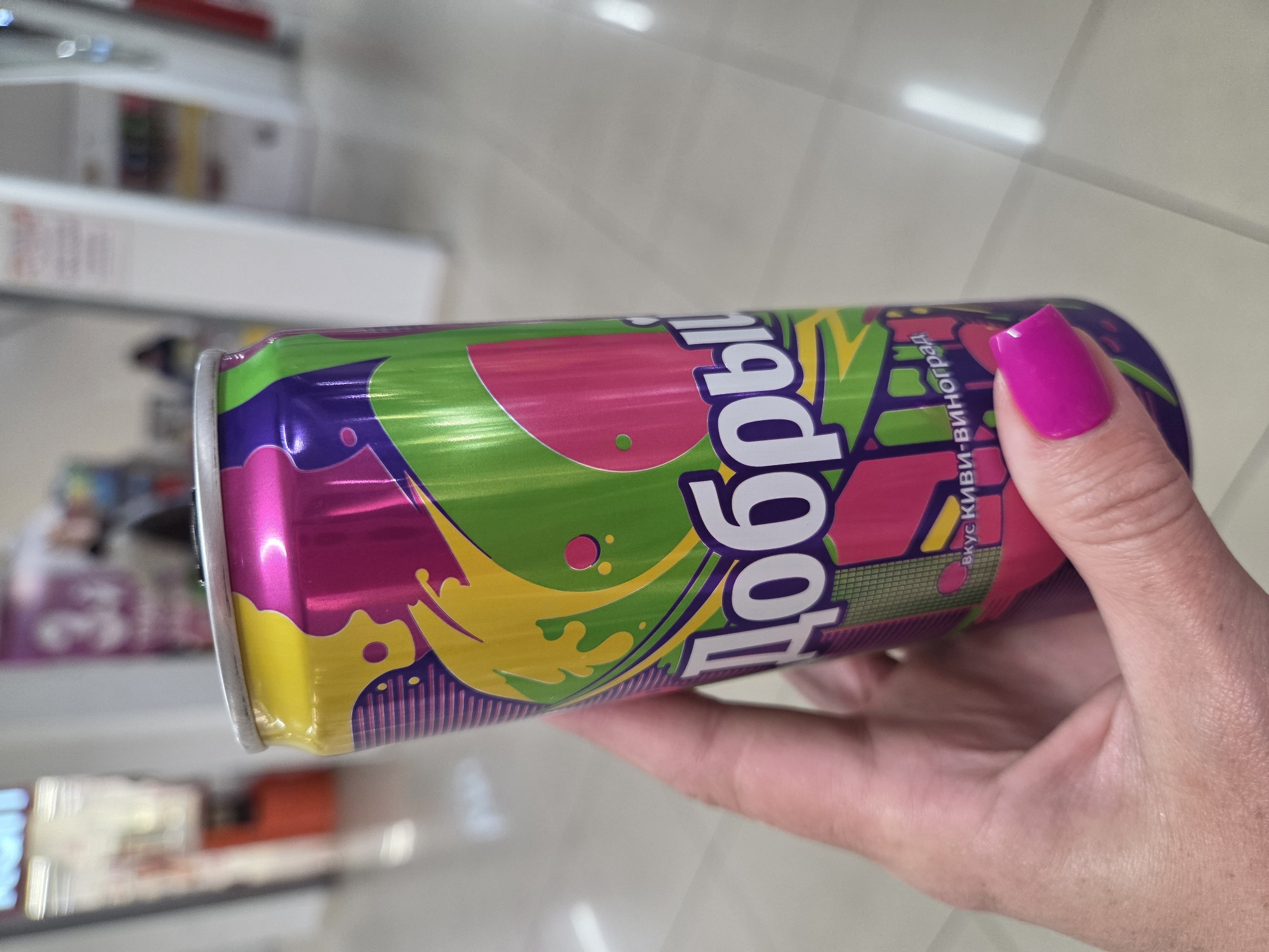 A hand with a bright pink fingernail is holding a can of Dobryy brand soda with a kiwi-grape flavor. The can is primarily purple, green, pink, and yellow. The background is blurred, but it appears to be a store shelf. The location is Birobidzhan, Russia.