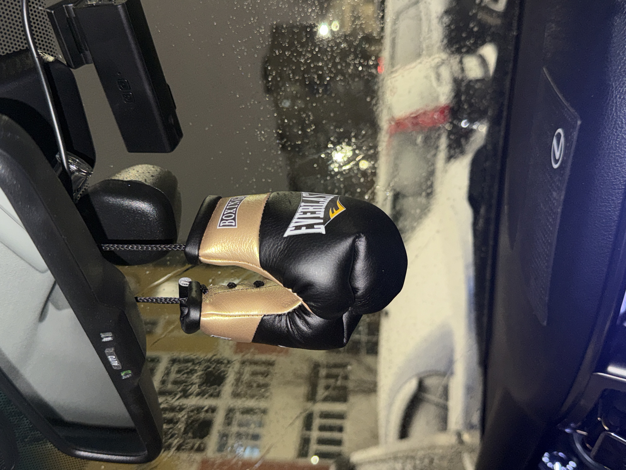 The image shows a pair of miniature Everlast boxing gloves hanging from the rearview mirror of a car. The gloves are black and gold. The car's windshield is covered in raindrops, and the view outside shows a blurry cityscape at night. Due to the lack of visual evidence, I cannot accurately determine if the location is in Nizhnevartovsk, Russia.