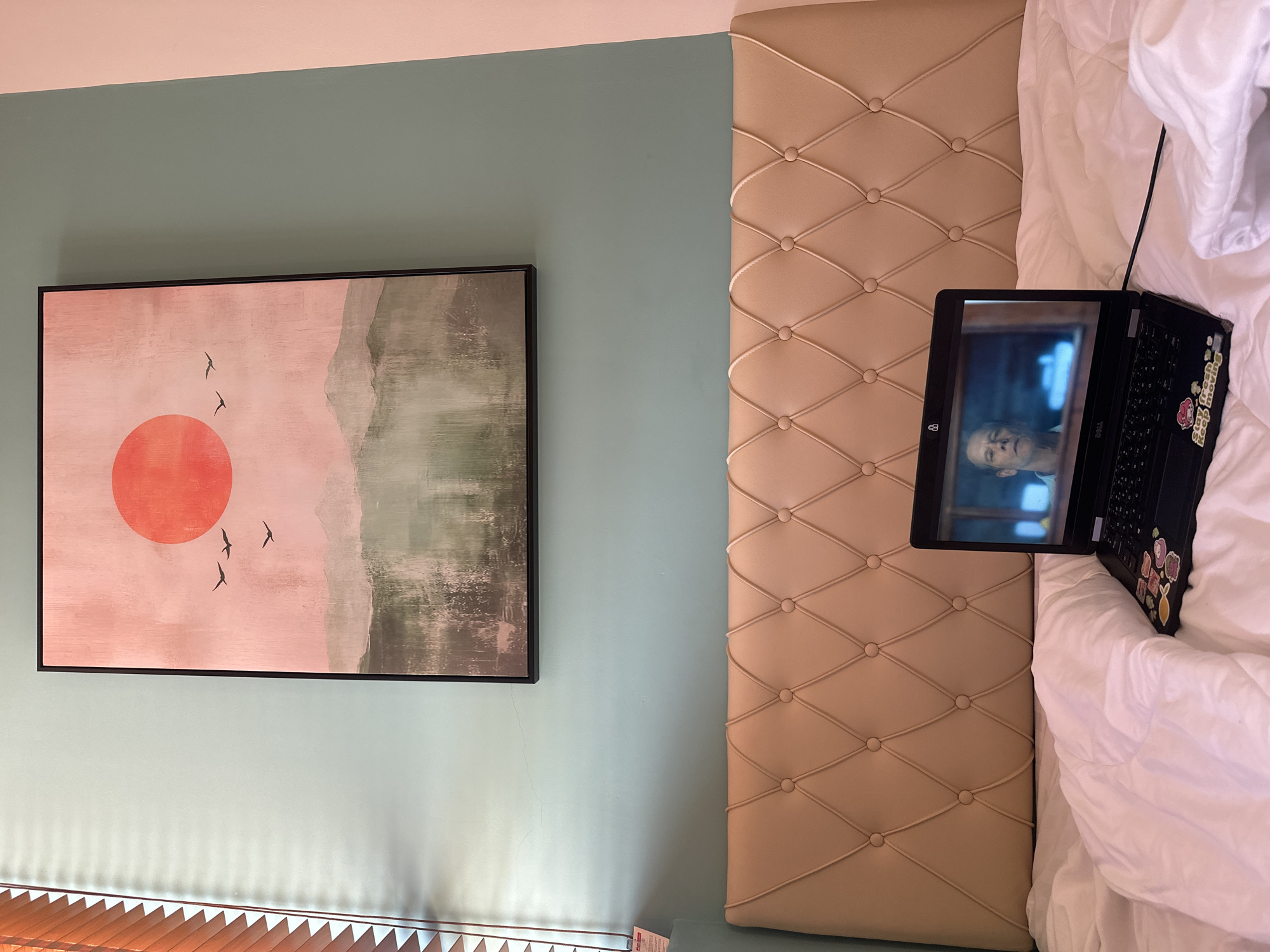 A laptop is open on a bed with white bedding in a room. The laptop screen displays a person's face, and the laptop itself has several stickers on it. Behind the bed is a tufted headboard. Above the headboard is a painting on a light blue wall. The painting depicts a red sun and silhouetted birds over a landscape of mountains. To the left of the painting, wooden blinds are visible.