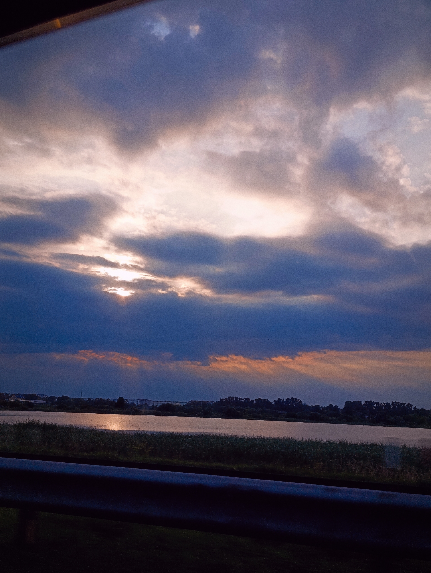 A dramatic sky with dark, billowing clouds is illuminated by shafts of sunlight breaking through. Below the sky, a calm body of water reflects the light, with a dark line of vegetation along its edge. In the distance, a silhouette of trees and buildings is visible along the horizon. The foreground is dominated by a dark, metallic barrier, possibly a guardrail, casting a shadow. The overall impression is one of a serene landscape at dusk.