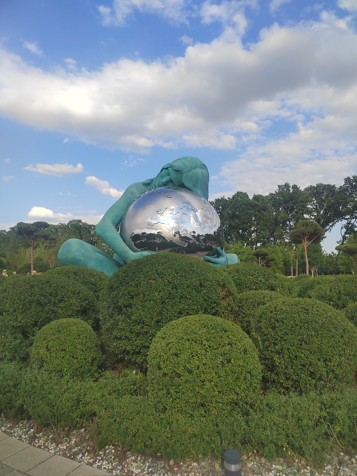 The photograph captures a striking sculpture of a giantess cradling a mirrored sphere, presumably representing the Earth, in Vityazevo, Russia. The sculpture, with its turquoise patina, is partially obscured by meticulously manicured, rounded green bushes arranged in a tiered landscape. In the background, a dense line of mature trees forms a natural backdrop under a bright blue sky dotted with fluffy white clouds. The mirrored sphere reflects the sky and surrounding greenery, adding a dynamic and almost ethereal quality to the scene. In the distance, several figures, appearing small in comparison to the sculpture, are visible amidst the trees, suggesting a park or public garden setting. A paved walkway and a light-colored gravel path are visible in the foreground, hinting at visitor access and landscaping.