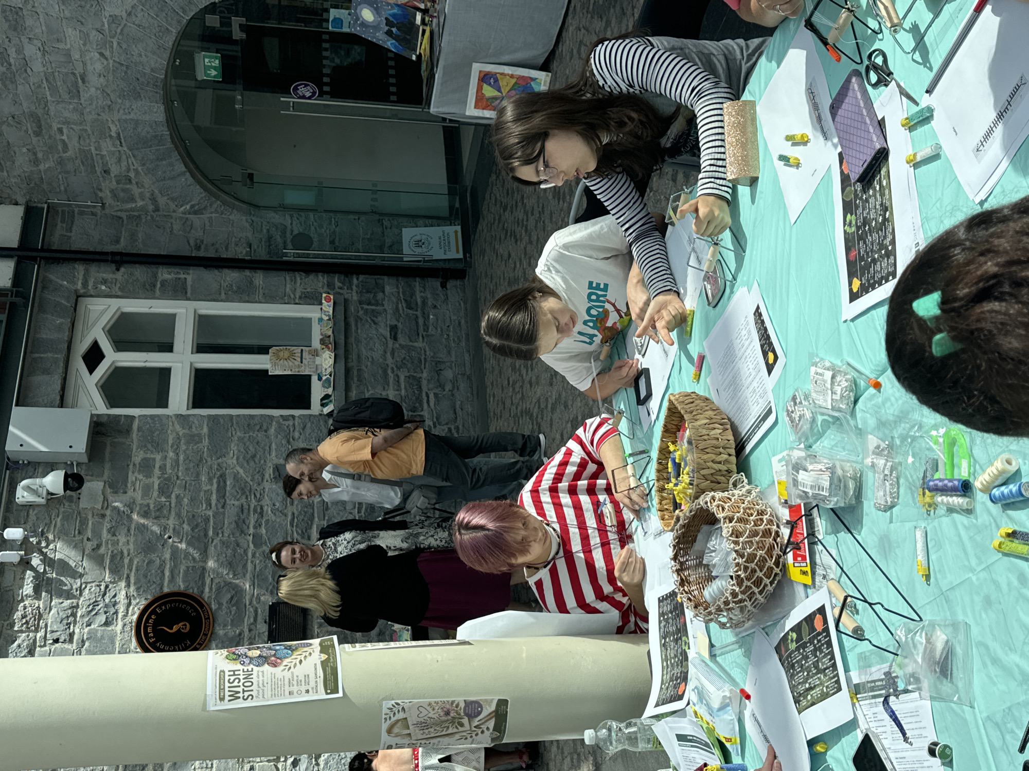 This image depicts an outdoor event taking place in Kilkenny, Ireland, likely at or near the "Kilkenny Famine Experience" which is indicated by a sign on a stone building in the background. The scene is set on a sunny day, with a stone building forming the backdrop, featuring an arched entrance with glass doors.
In the foreground, a turquoise tablecloth covers a table where several individuals are engaged in crafting or workshop activities. Three young women are seated at the table, focused on their work. One person, wearing a red and white striped shirt, is concentrating on something in front of them, holding a thin tool. Another young woman, in a white t-shirt with lettering, has her hands on the table and appears to be explaining or demonstrating something. The third young woman, wearing a striped long-sleeved shirt and glasses, is actively gesturing with her hands as if speaking or instructing.
The table is cluttered with various crafting supplies, including spools of thread, small containers of beads or glitter, paper instructions with diagrams, scissors, and small baskets. Bags of small items and tools are also visible.
In the middle ground, a group of adults are standing and conversing. One woman in a black jacket and dark skirt is talking to another woman in a white shirt and a man in an orange t-shirt. Behind them, more people can be seen standing.
Posters are displayed on a pillar to the left of the scene, one of which advertises a "WISH STONE" event. The overall atmosphere suggests a community gathering or an educational workshop, possibly related to crafts or heritage. The stone architecture and the sign for the "Kilkenny Famine Experience" firmly place the location in Kilkenny, Ireland.