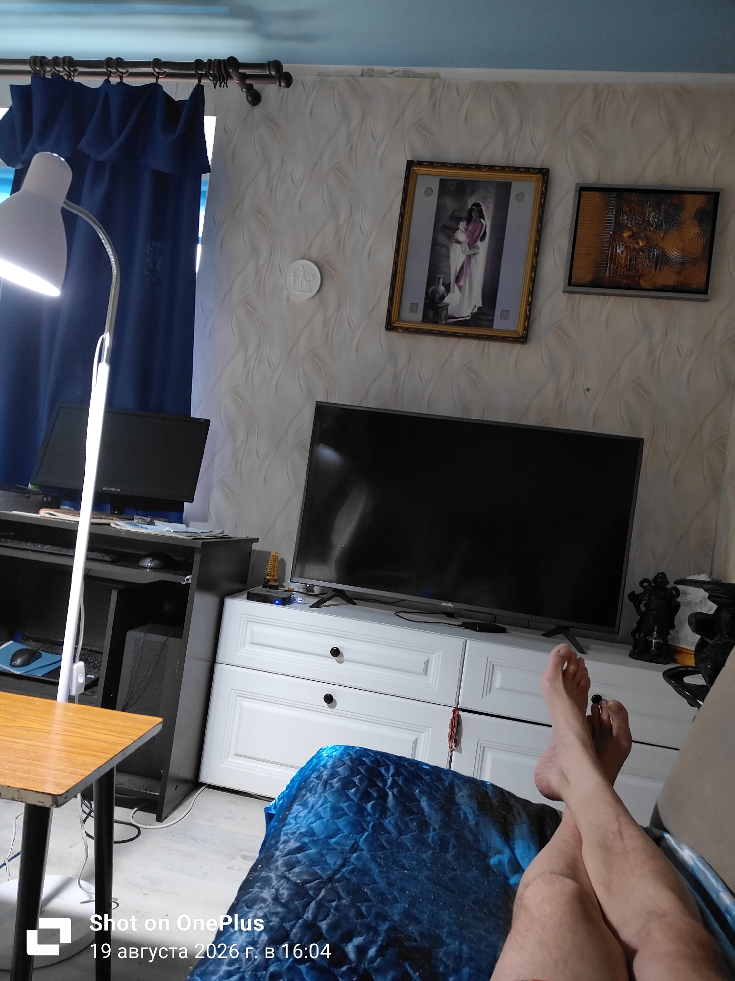 The image depicts an indoor scene, likely a living room or study, in Svetly, Russia. A desk with two computer monitors and a computer tower is visible on the left. A floor lamp with a white shade illuminates the area. On the wall above the desk, there are two framed pictures, one depicting a woman in a white dress and the other an abstract design. A large flat-screen television sits on top of a white dresser with multiple drawers. In the foreground, a person's legs and feet are crossed, resting on a blue quilted blanket. The timestamp indicates the photo was taken on August 19, 2026, at 16:04.