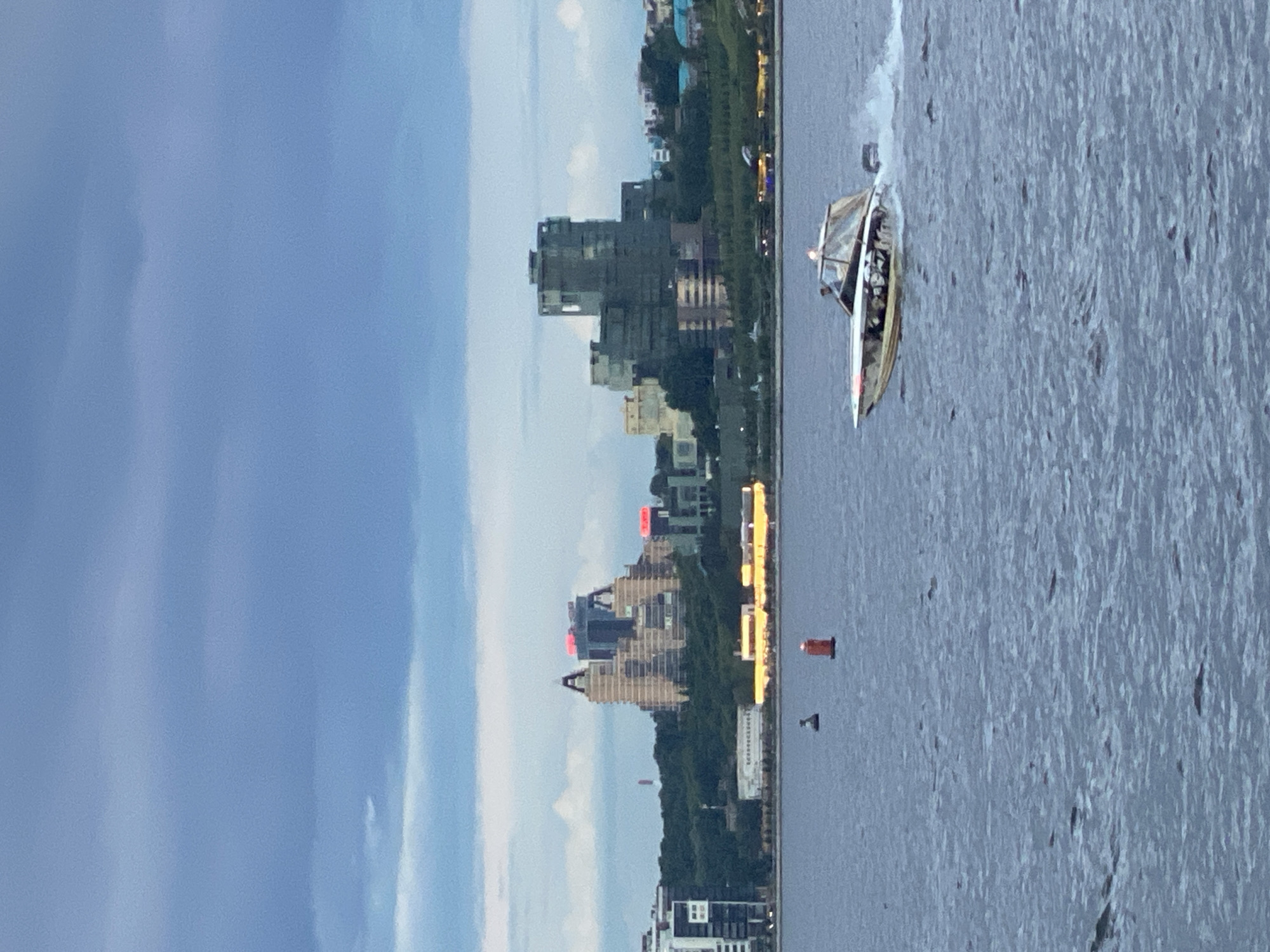 The image, correctly oriented, depicts a panoramic view of a city skyline along a wide body of water under a late afternoon or early evening sky. The setting is likely Kazan, Russia, given the context.
In the foreground and mid-ground, a broad expanse of grey-blue water stretches across the frame. A sleek, white and silver motorboat is actively moving across the water from right to left, leaving a prominent white wake behind it. The boat has a cabin structure and appears to carry passengers, though individual details are indistinct due to distance. To the left of the boat, two buoys are visible in the water: a bright orange cylindrical buoy and a smaller, dark, conical buoy located further to its left.
The far bank features a dense cityscape, with a variety of buildings rising behind a strip of green trees. The architecture includes modern high-rises and distinctive, more complex structures. Several buildings have lights illuminated, and some taller ones exhibit notable red lights on their rooftops, indicating the transitional time of day between daylight and night. The sky overhead is a soft, muted blue with subtle cloud formations, providing even, diffused lighting across the scene. The overall atmosphere is calm and serene, showcasing urban life against a natural waterscape. No discernible text is visible on any objects or buildings.