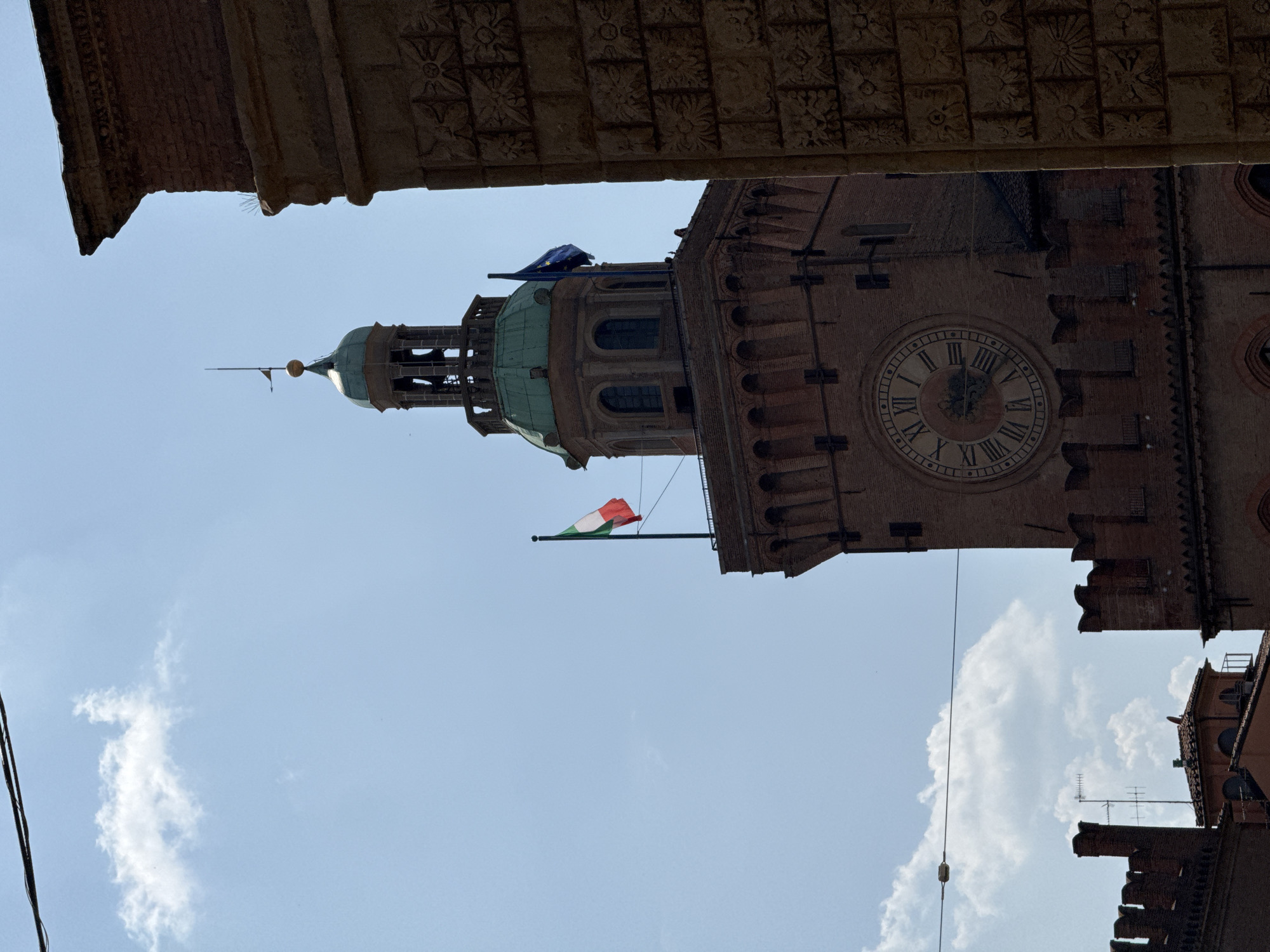 A clock tower stands prominently against a bright blue sky with scattered clouds. The tower is constructed of red brick and features a large circular clock face with Roman numerals. Above the clock, the Italian flag is visible flying from a flagpole, and next to it, the European Union flag is also displayed. The tower is topped with a green dome and a smaller spire. To the right of the clock tower, a portion of a building with ornate stone carvings is visible. In the lower left of the image, other red brick structures with crenellated tops can be seen, suggesting an architectural style common in Bologna, Italy. The overall scene captures a historical and architectural landmark in an urban environment.