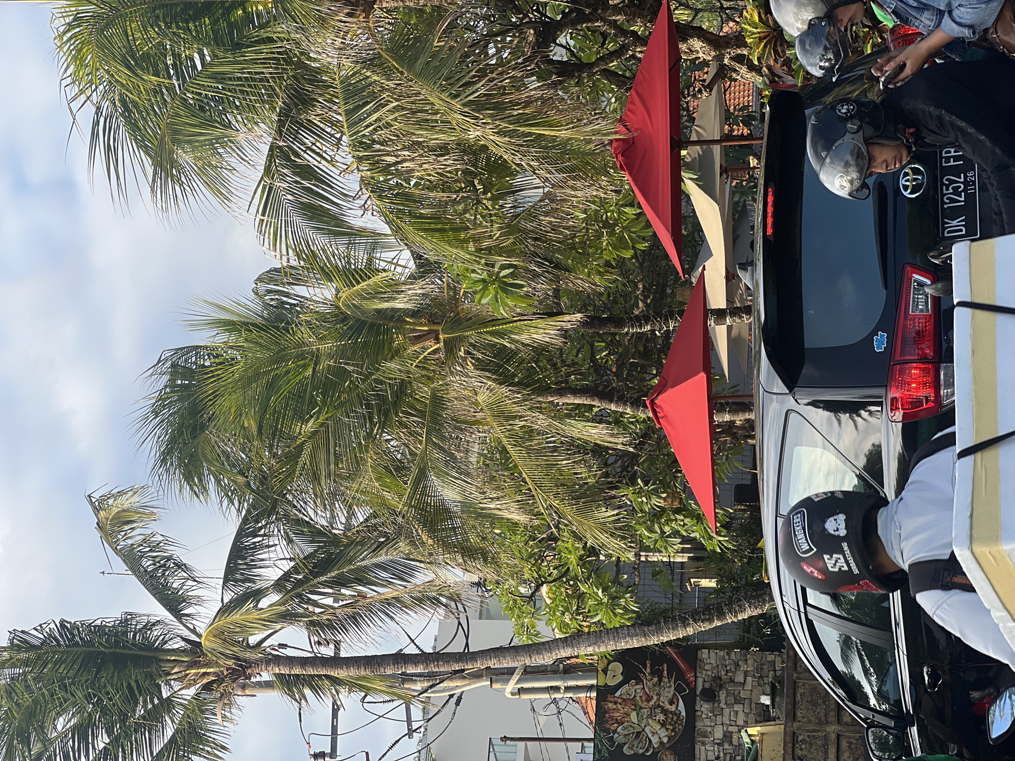 In Kuta, Indonesia, a street scene unfolds with tropical vegetation and vehicles. Palm trees with lush fronds dominate the upper portion of the image, their leaves rustling against a sky with scattered clouds. Below the trees, a black SUV is parked, its rear visible. A person wearing a black helmet with stickers, including one that says "WANGKERS," sits on a motorbike behind the SUV. Another individual wearing a helmet is seen in the right side of the image, also near the SUV. Red umbrellas are positioned above outdoor seating areas, suggesting a restaurant or cafe. A sign with an image of seafood is attached to a stone wall on the left side of the frame. Power lines and poles are visible amidst the foliage, indicating an urban setting. The overall impression is a snapshot of daily life in a tropical, coastal city.