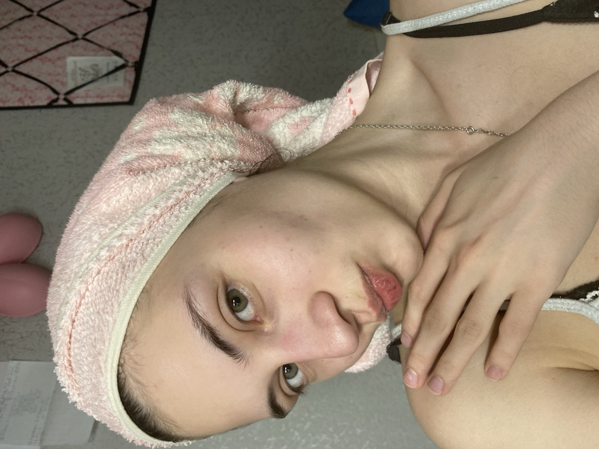 A person is pictured indoors, angled in the frame, with a pink and white towel wrapped around their head. They have fair skin, dark eyebrows, and green eyes. A silver chain is visible around their neck, and part of a black and white garment is seen near the shoulder. The person's hand is positioned near their face. A pink object and a basket are visible in the background, along with a few sheets of paper. Location information is not discernible from the image.