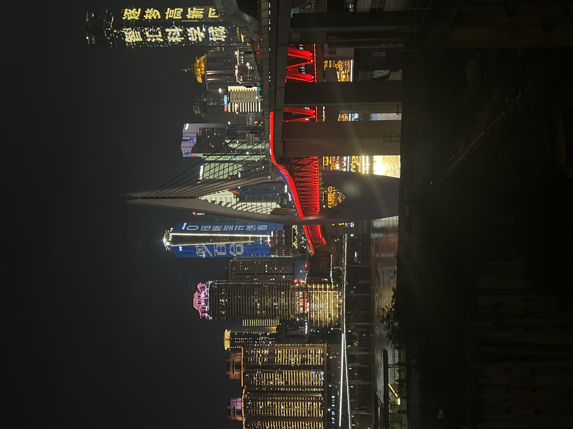 The image depicts a night scene in Jiangbei Cheng Street, China, showcasing a modern cityscape dominated by illuminated skyscrapers and a prominent cable-stayed bridge. The bridge, a central element, features a white pylon and a red illuminated deck that spans across a body of water, reflecting the city lights. Several tall buildings, varying in architectural style and illuminated with a mix of yellow, pink, and blue lights, rise behind the bridge. One skyscraper on the right prominently displays yellow characters, possibly a company name or slogan. The lower part of the image shows a dark foreground, obscuring details of the immediate surroundings, but the water below the bridge is visible, rippling with reflected light. No people are discernible in the frame.