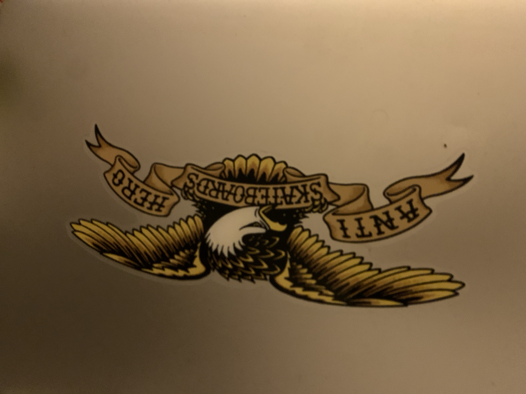 The image shows a sticker featuring an eagle design with a banner that reads "SKATEBOARDS" in the center, "HERO" on the left side, and "ANTI" on the right side. The eagle has a white head and detailed feathers. The overall color scheme is gold, black, and white. The background appears to be a plain, light-colored surface. There is no indication that the image was taken in Barcelona, Spain.