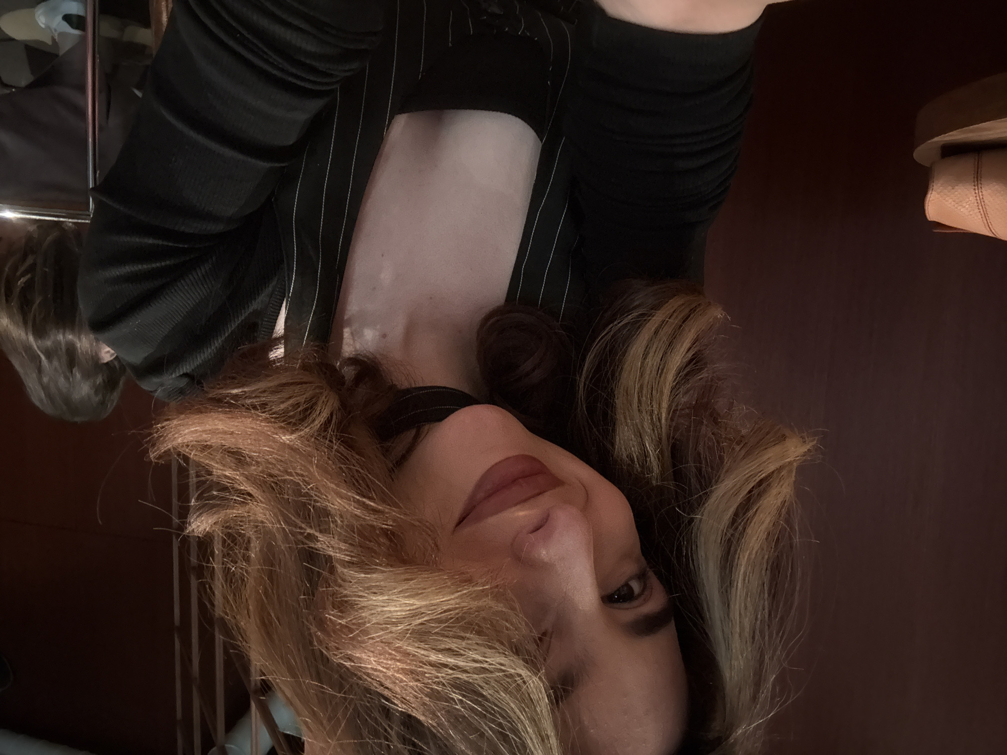 A close-up, portrait-oriented shot features a woman looking towards the camera with a subtle smile. Her head is tilted, and her voluminous, highlighted brown hair with blonde streaks frames her face and cascades around her shoulders. She wears a black ribbed top and a dark, possibly black, pinstriped blazer or jacket over it. A thin black choker-style accessory is visible around her neck. Her makeup includes lipstick and defined eyebrows.
The background is primarily dark, with rich brown tones suggesting a wooden surface or wall. To the left, a reflective surface, possibly a mirror or polished metal, shows a blurred reflection of dark hair, suggesting another person or a different angle of her own reflection. To the far right, a light brown object with visible texture, resembling a leather bag or shoe, rests on a dark surface. The lighting is soft, suggesting an indoor setting with ambient or artificial light. No visible text is present in the image. The location context provided is Yerevan, Armenia, though no specific visual cues confirm this within the image itself.