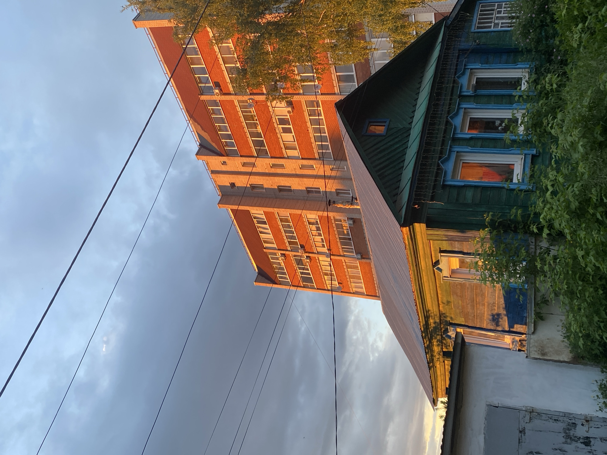 The image shows a high-rise red brick building adjacent to a low, old wooden house. The wooden house has a green roof, blue window frames, and a weathered facade. A concrete structure with a door is connected to the wooden house. There are power lines crossing the sky. The photo was taken in Votkinsk, Russia.