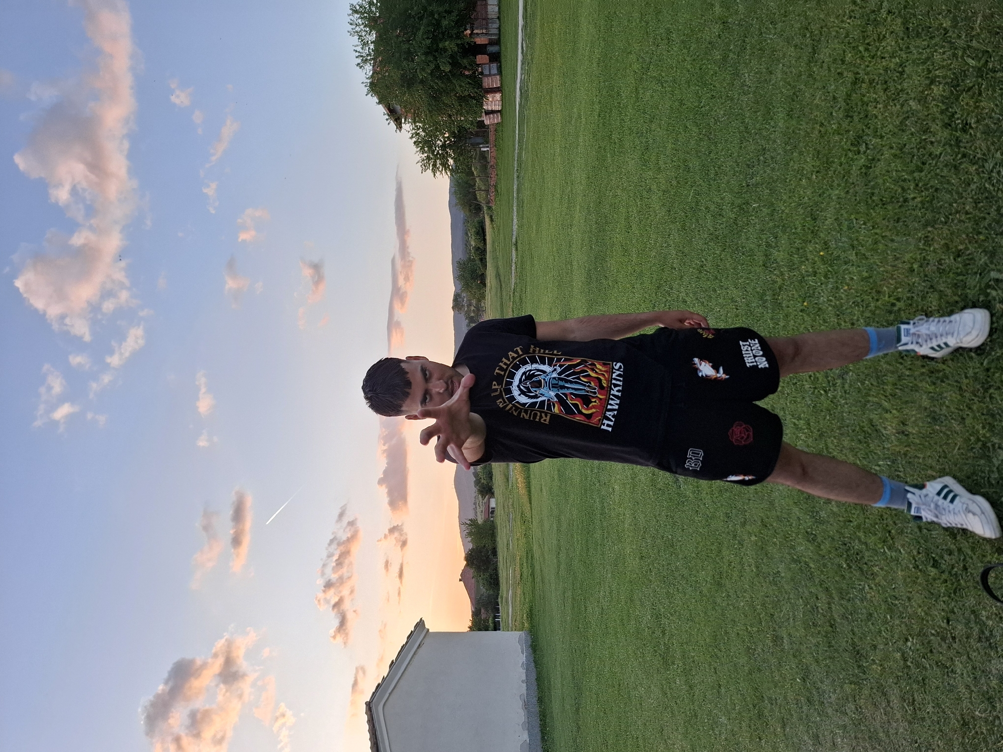 A young man stands in a grassy field in Krapets, Bulgaria. He wears a black T-shirt and shorts, and white sneakers with green stripes. The sky is a gradient of blue and pink, suggesting sunrise or sunset. A building is visible in the lower left corner. Trees and a distant mountain range are in the background. The man is reaching toward the camera with his right hand.
