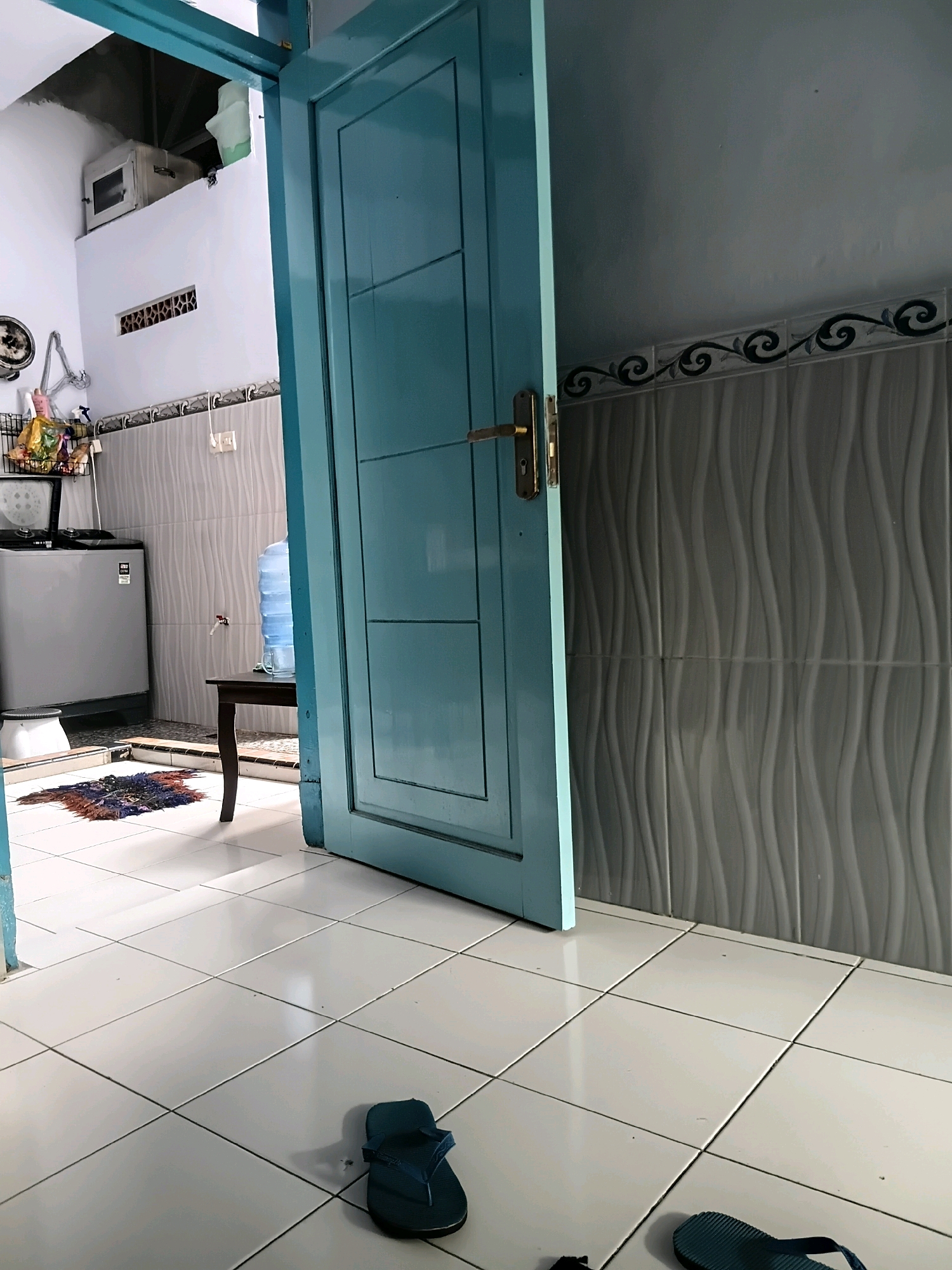 The image depicts the interior of a room, possibly a utility area or small kitchen, in Tanggul Wetan, Indonesia. The scene is dominated by a light blue door, partially ajar, which opens inwards. The flooring is covered in large, white square tiles. To the left, a portion of a silver washing machine is visible, with a small rug on the floor in front of it. Further left, a shelf unit holds various cleaning supplies. A tall water dispenser with a blue-tinted bottle sits on a dark wooden side table. The walls are tiled with a wavy pattern in a neutral grey color. Above the tiled walls, a decorative border with black and white scrollwork is present. The upper portion of the wall is painted white, with a rectangular ventilation grate visible. A small, silver appliance with a black window is mounted high on the wall above the grate. Two dark blue flip-flops are on the floor, one in the foreground to the right, and another partially visible in the lower right corner. The lighting suggests an indoor environment, possibly during the daytime, with shadows cast by the door and the side table.